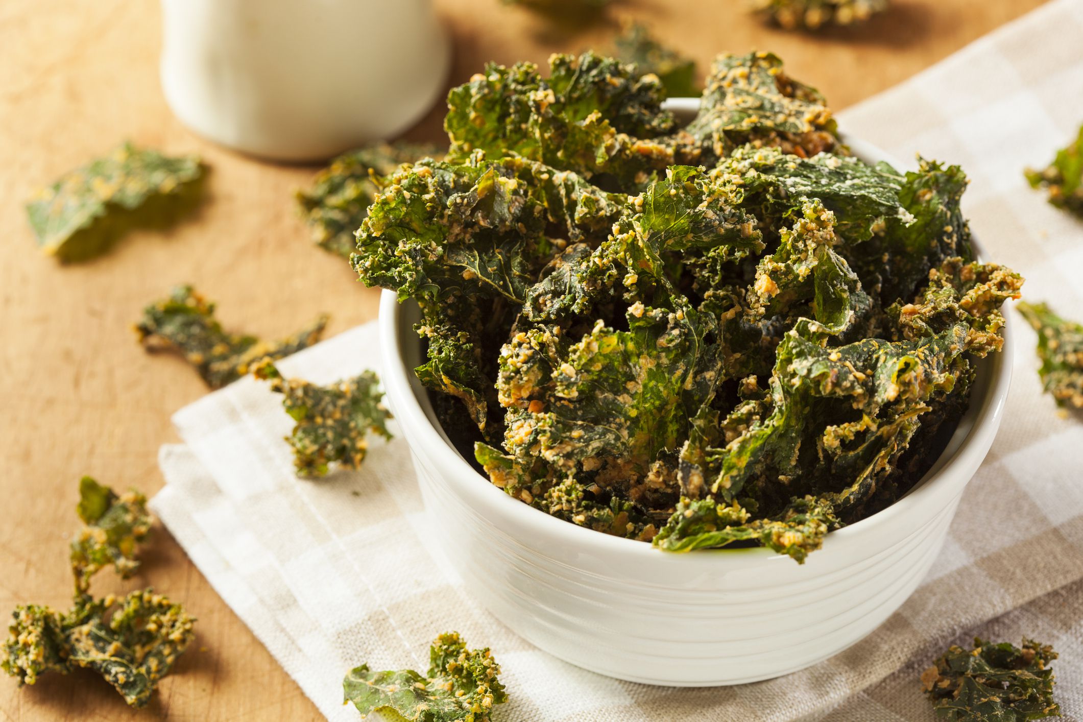 Top view of kale chips in a small white bowl on a white and tan checkered napkin surrounded by kale chips on a natural wooden table with a blurred background