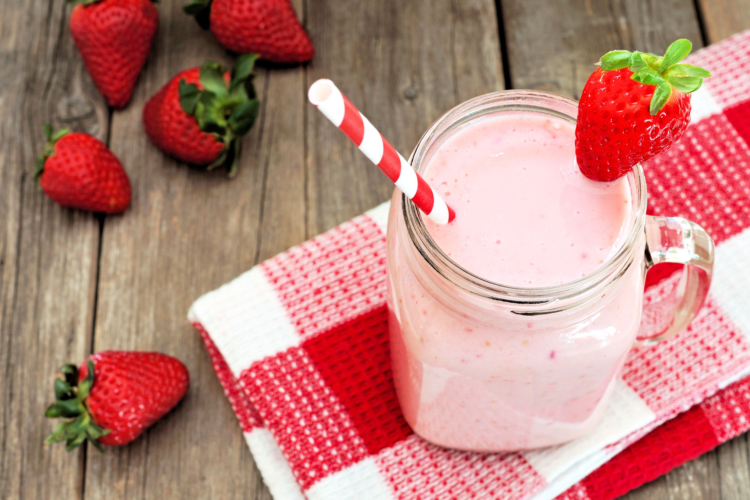Top view of strawberry smoothie in a glass mason jar on a red and white checkered napkin with strawberries on a wooden table