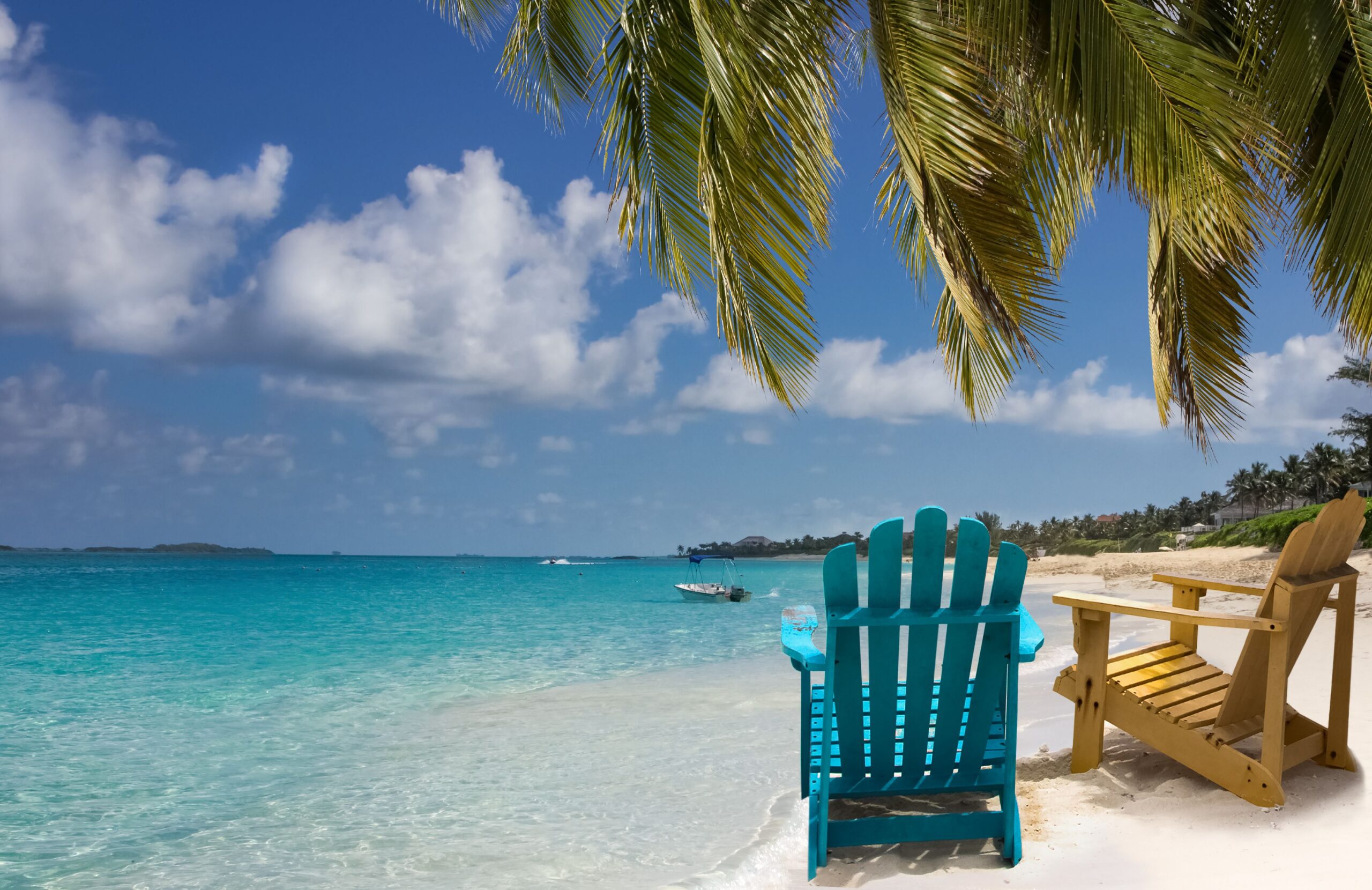 Focus on two empty Adirondack chairs on the white sandy beach with ocean in Nassau, Bahamas, as seen through palm tree leaves on the top