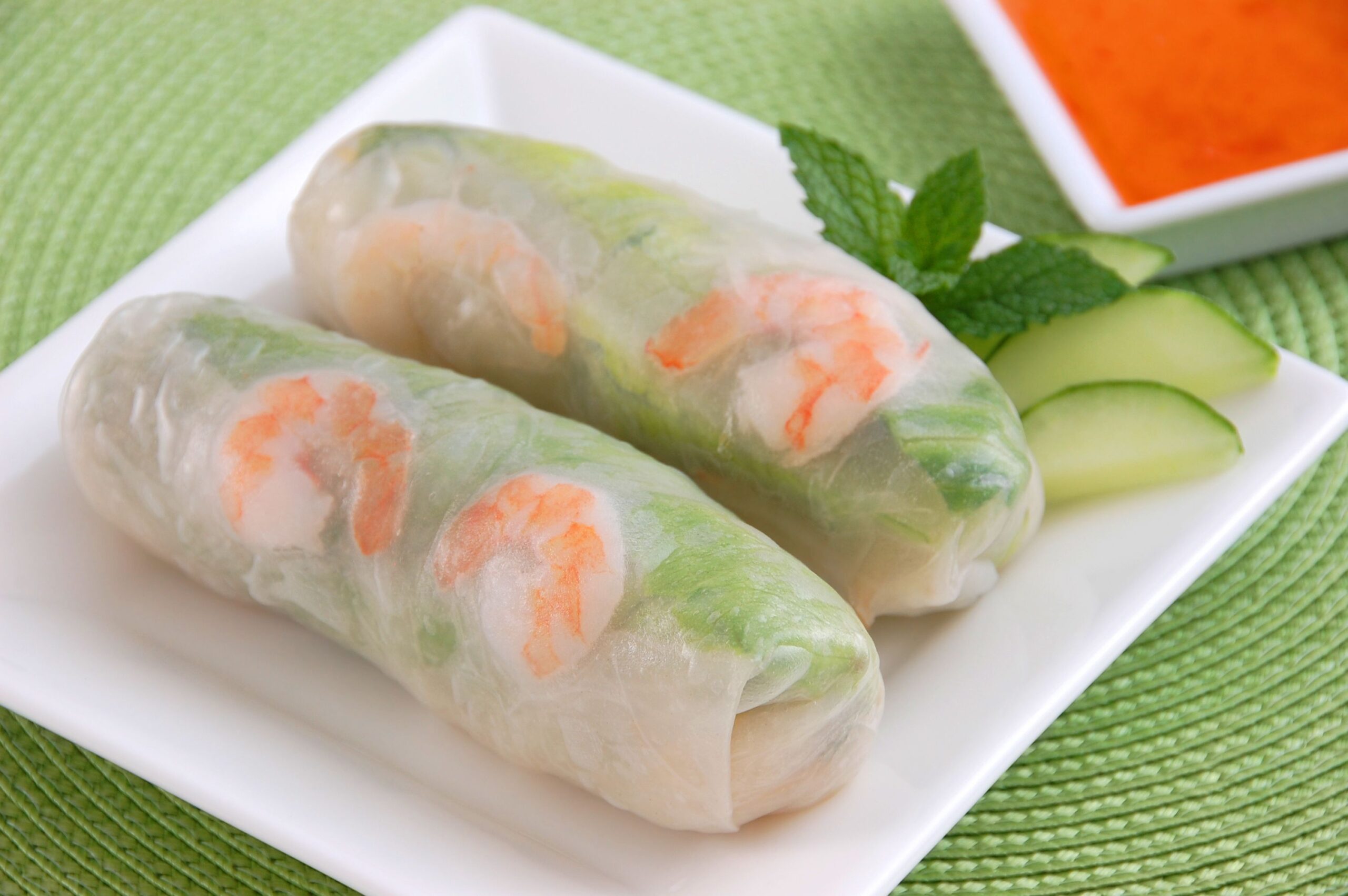 Two shrimp and mango summer dipping rolls on a square white plate on a round avocado green tablemat with a square bowl of orange dipping sauce