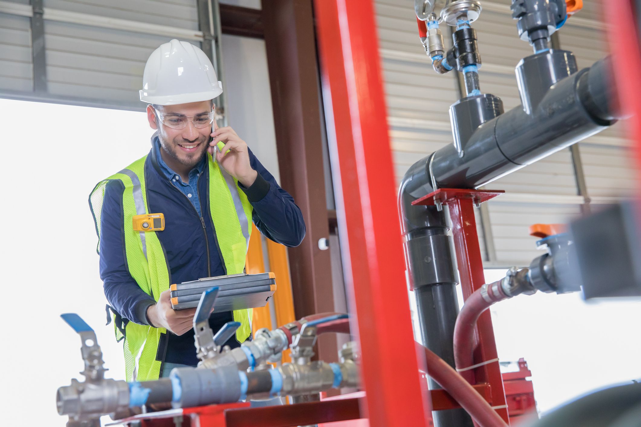 Young adult Hispanic man is an engineer working in oil and gas industry. He is inspecting pipeline equipment and making a phone call. Man is wearing hard hat and safety vest.