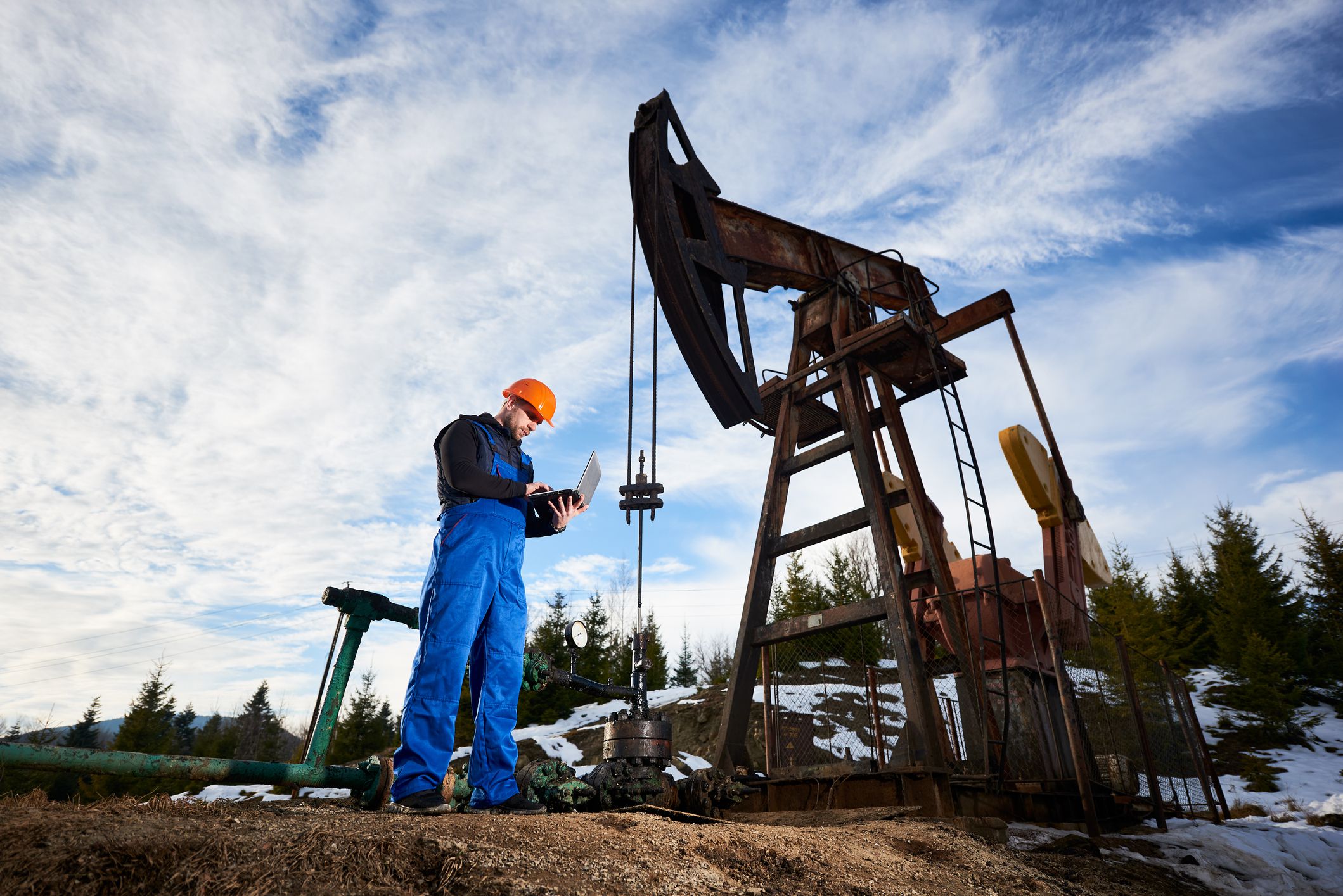 Oil worker using laptop, controlling work of petroleum pump jack. Drill operator in work overalls and helmet near oil pumping unit under cloudy sky. Concept of petroleum industry and oil extraction.