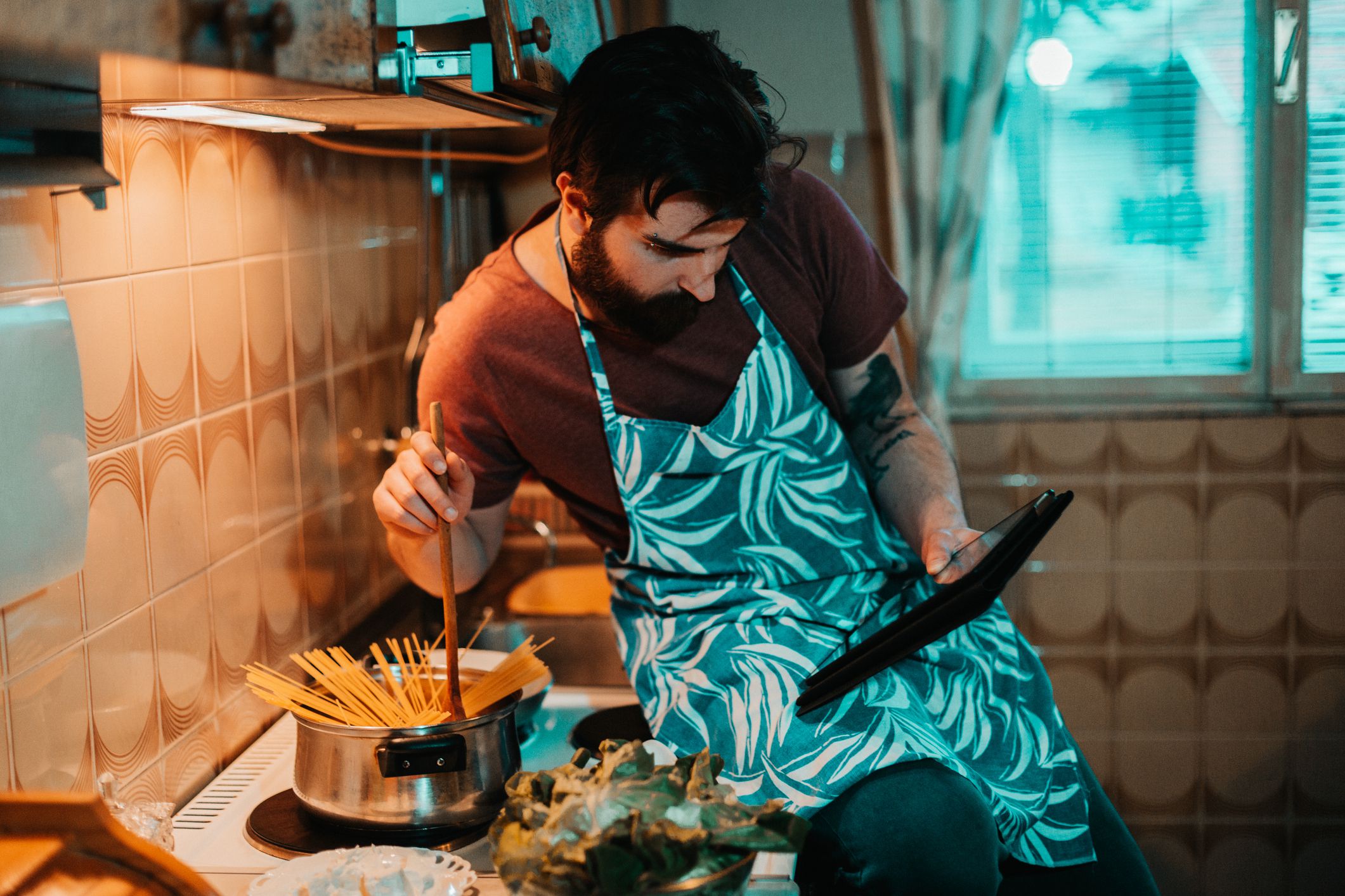 Handsome bearded man following recipe on digital tablet and preparing food in the kitchen at home