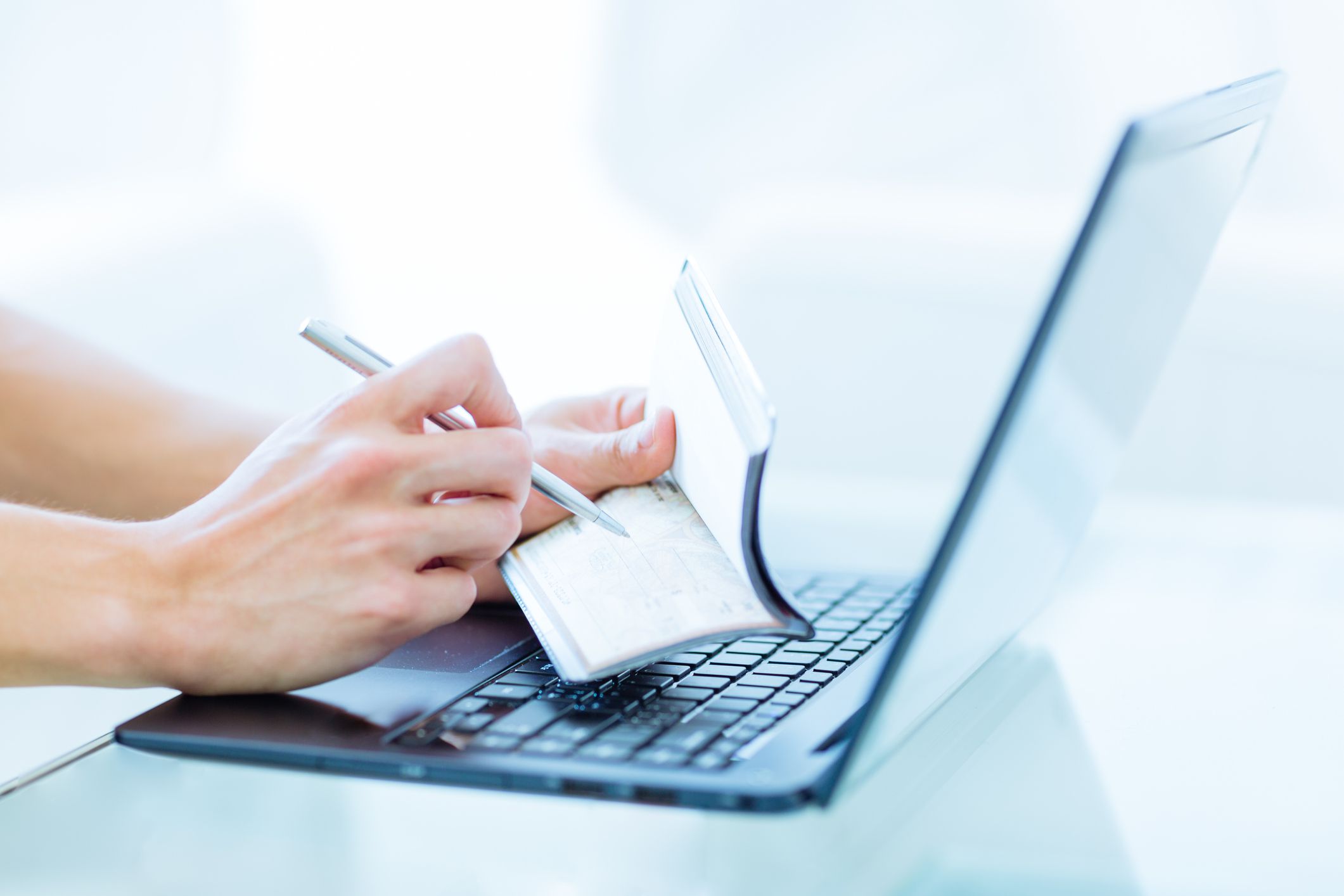 Closeup of a caucasian male adult writing on an empty personal chequebook while using a laptop computer in a modern environment.