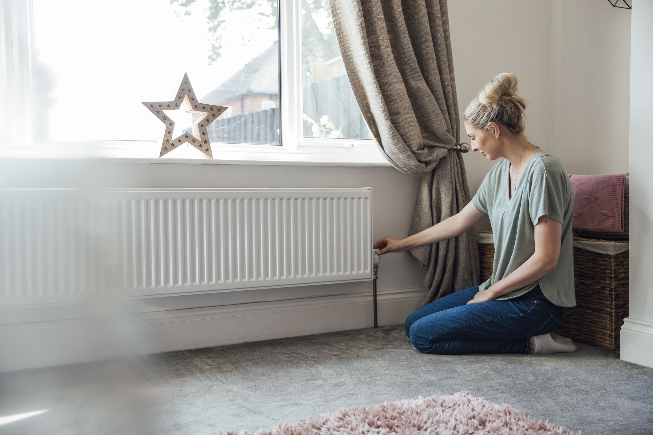 Side view of a young woman kneeling on the living room floor, adjusting the thermostat on the radiator.