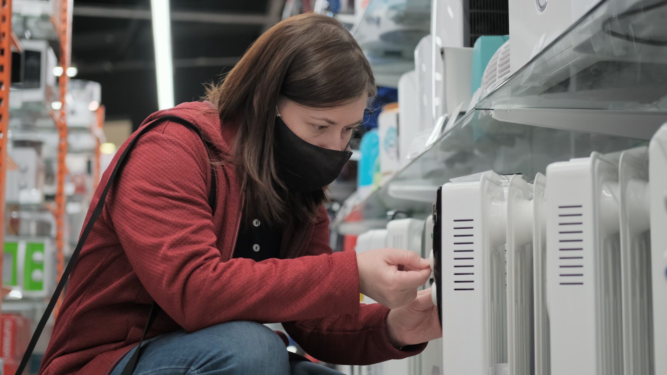 Caucasian a women wearing antivirus medical mask and red jacket picks out portable heater in electronics shop. Going to showroom during pandemic