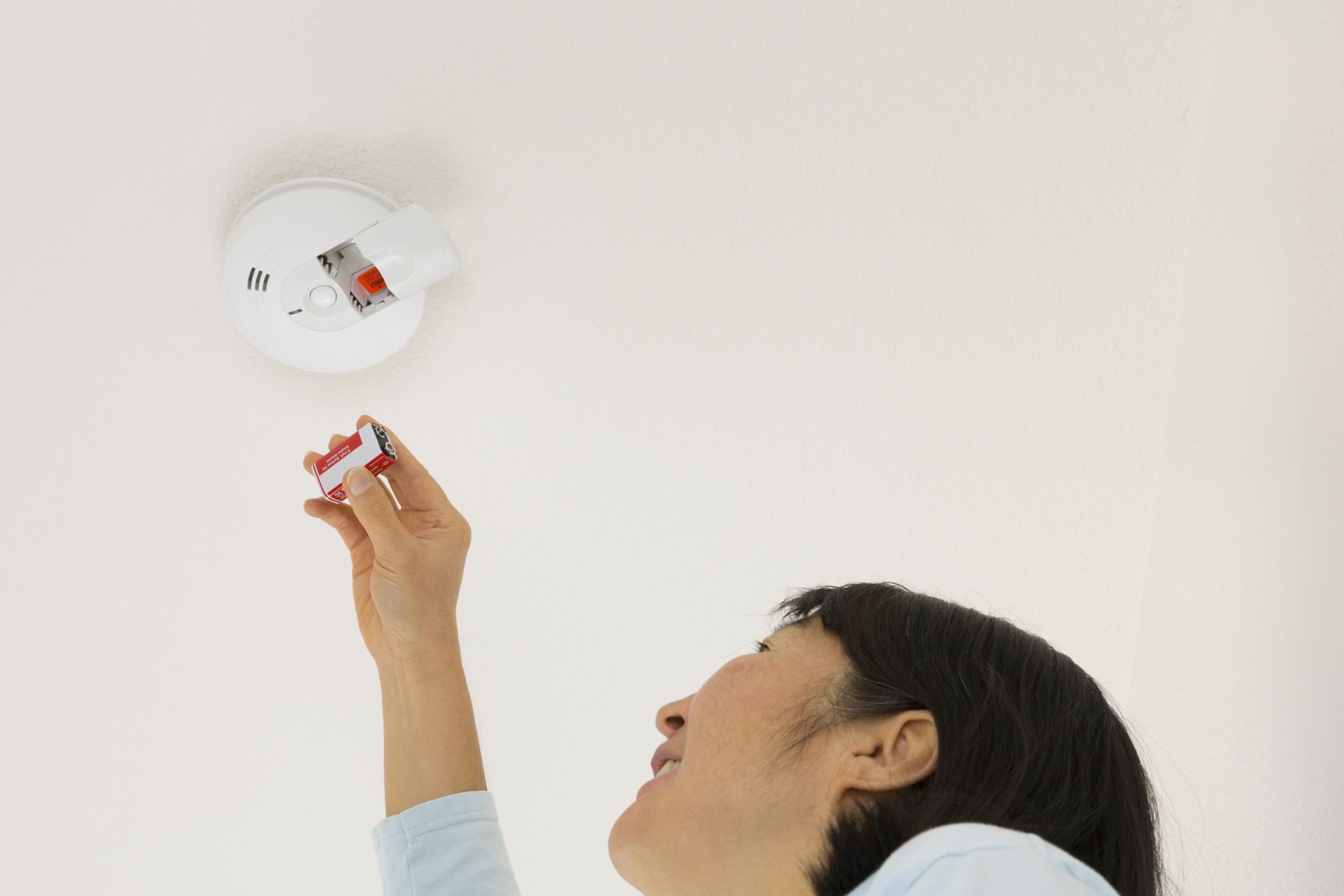 A middle-aged Asian woman inserting a 9V battery into a ceiling-mounted smoke detector.