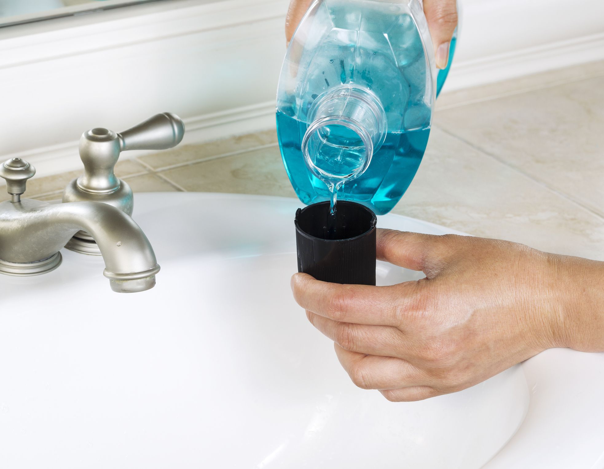 Horizontal photo of female hands putting mouth wash into black cap with white bathroom sink and faucet in background