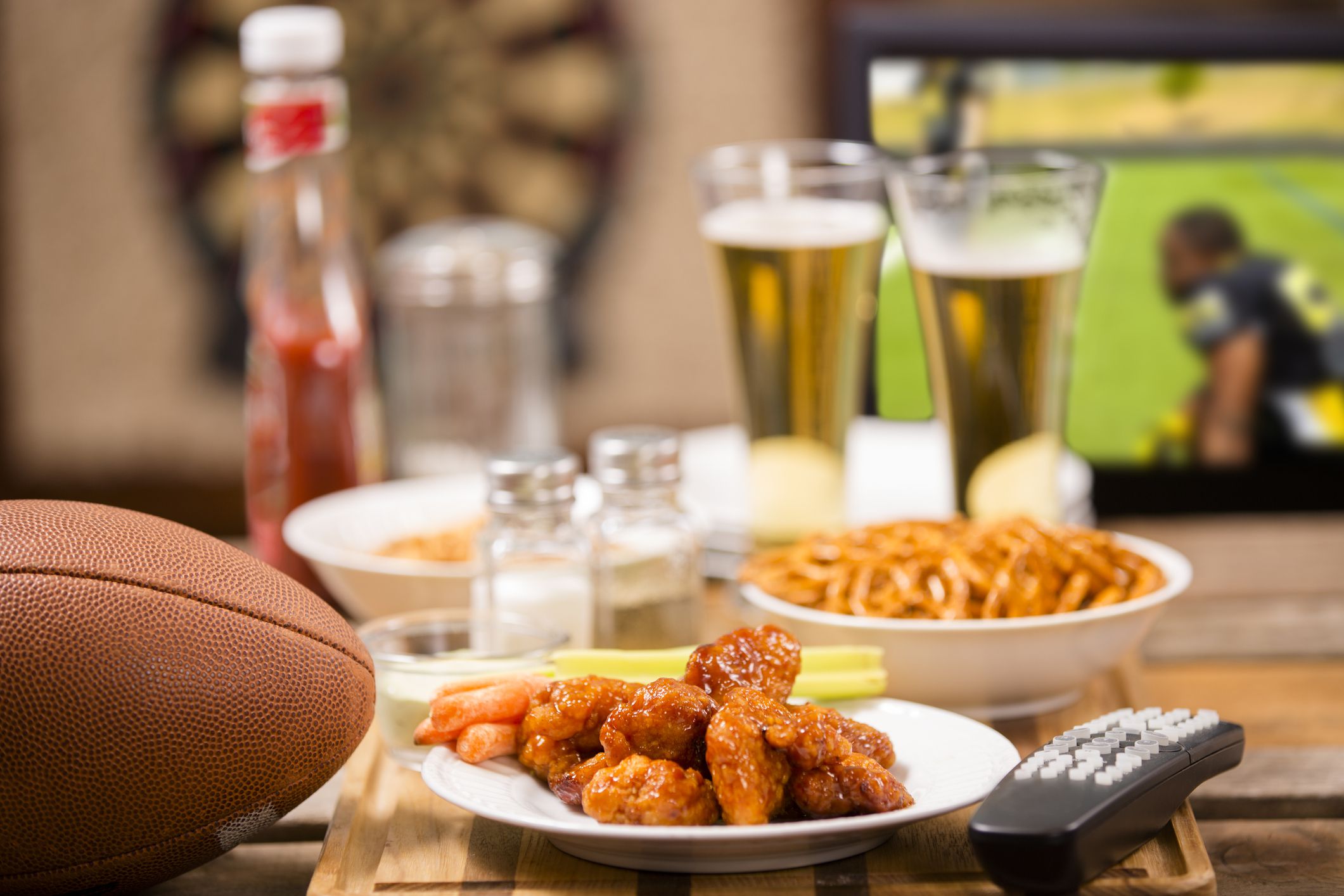 Hot wings and football in foreground. Beer in mugs in background with television. Football game on TV in a local pub or sports bar. Dartboard in background. Bar top. championship game party!