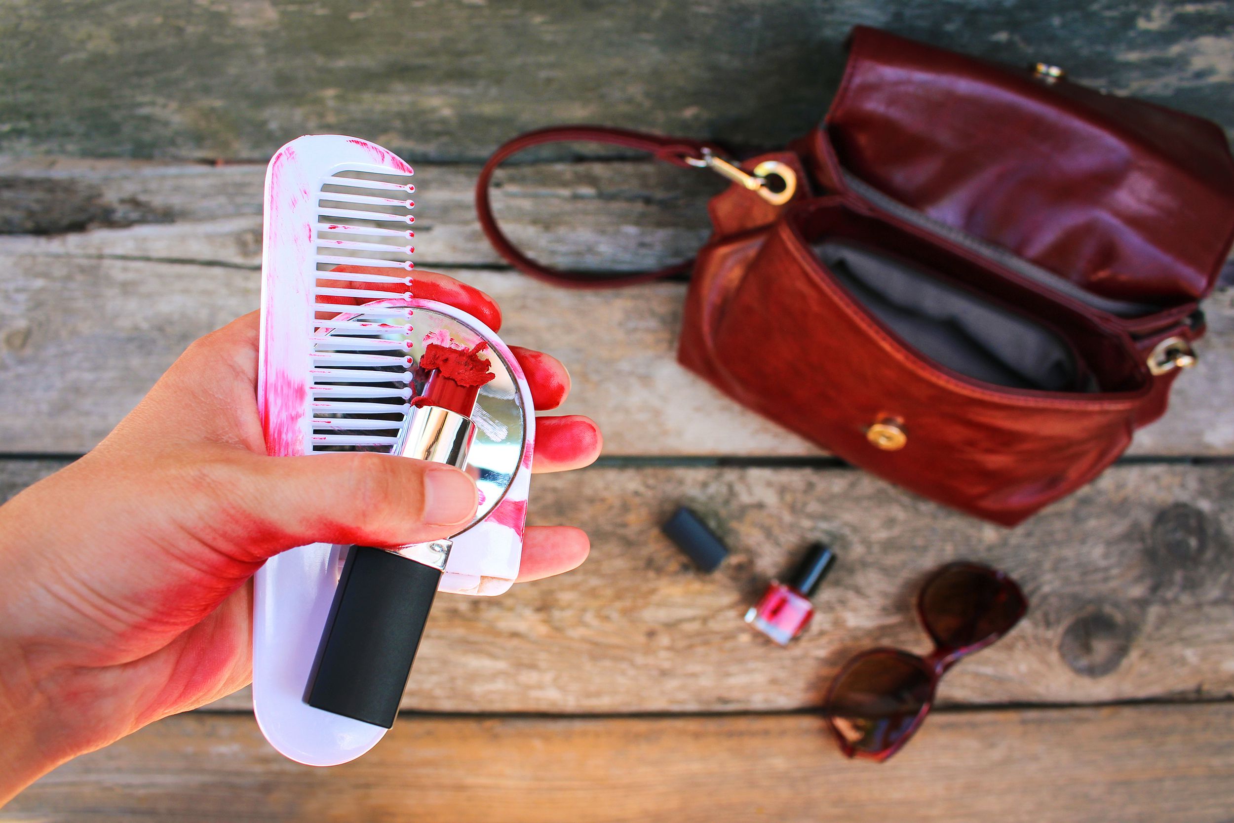 broken red lipstick, mirror, comb in women hand with purse below