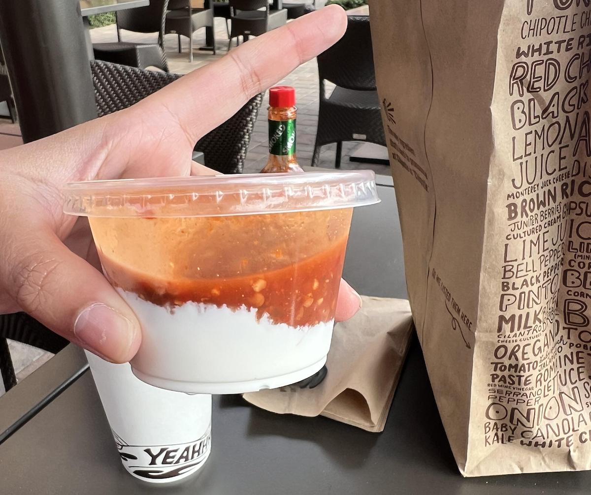 A hand holds a clear plastic cup with white sour cream topped with red salsa. In the background are a Chipotle paper bag, a drink cup, and a bottle of Tabasco sauce on a table with outdoor seating.