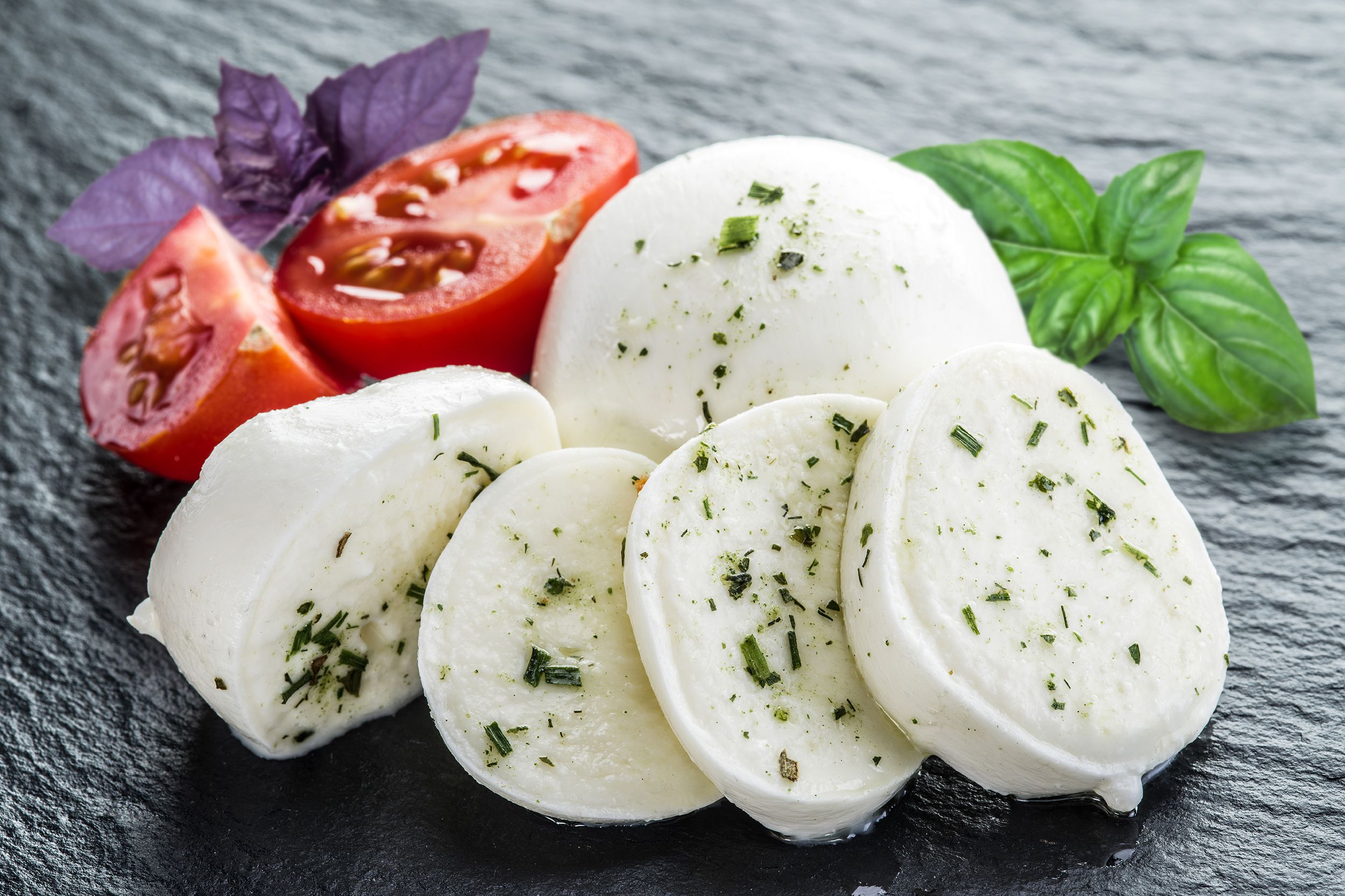 Closeup of five large slices of mozzarella with a cut tomato and a few herbs on a slab of slate