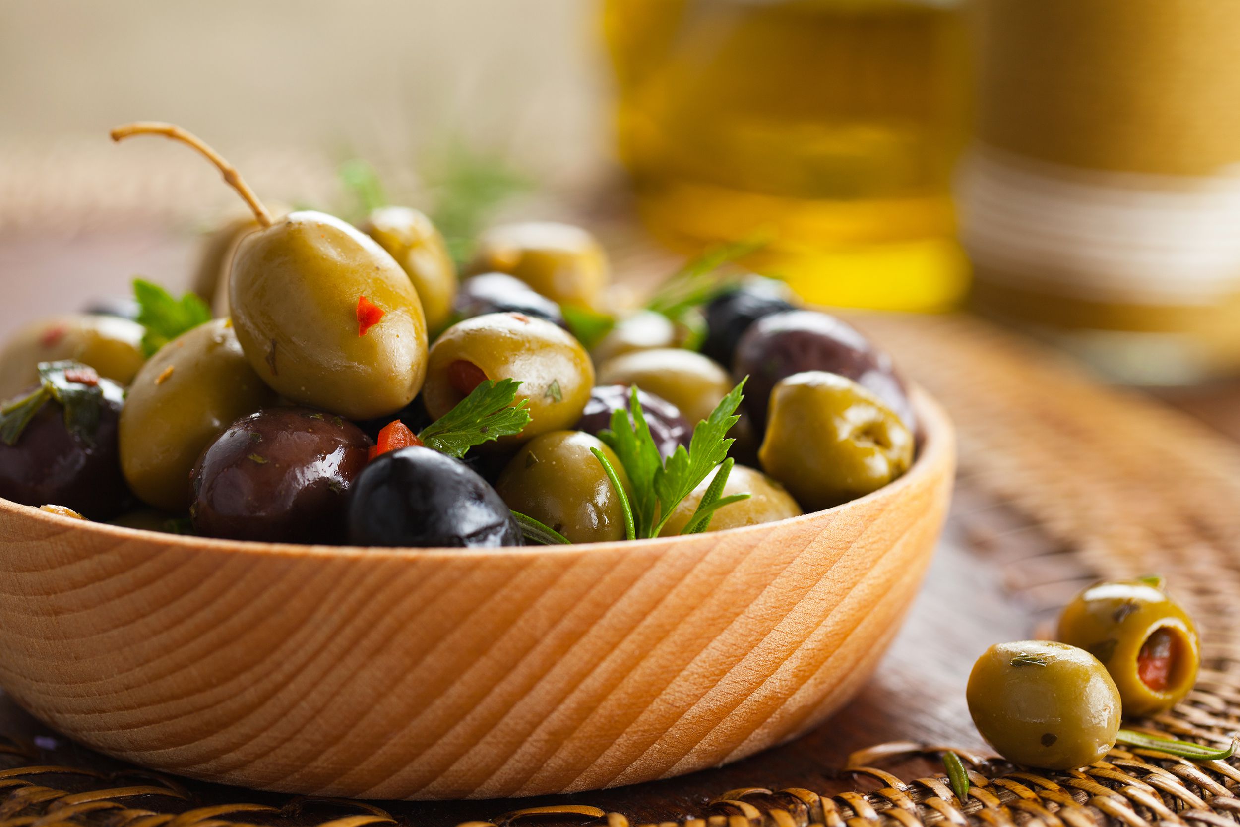 Closeup of marinated green, kalamata, and black olives in a wooden bowl on a brown basket weave placemat with a blurred background of olive oil