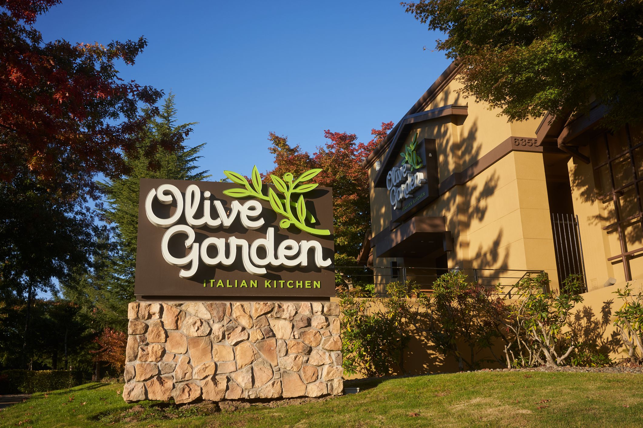 Sign in the foreground with garden area and with exterior in background of an Olive Garden restaurant, Lake Oswego, Oregon with a blue sky in the background during late summer