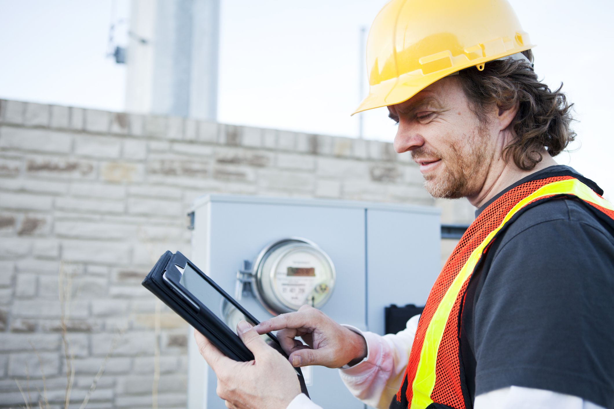 Male power line technician holding tablet to record electrical meter, looking at tablet, electrical power meter is in the background attached to building