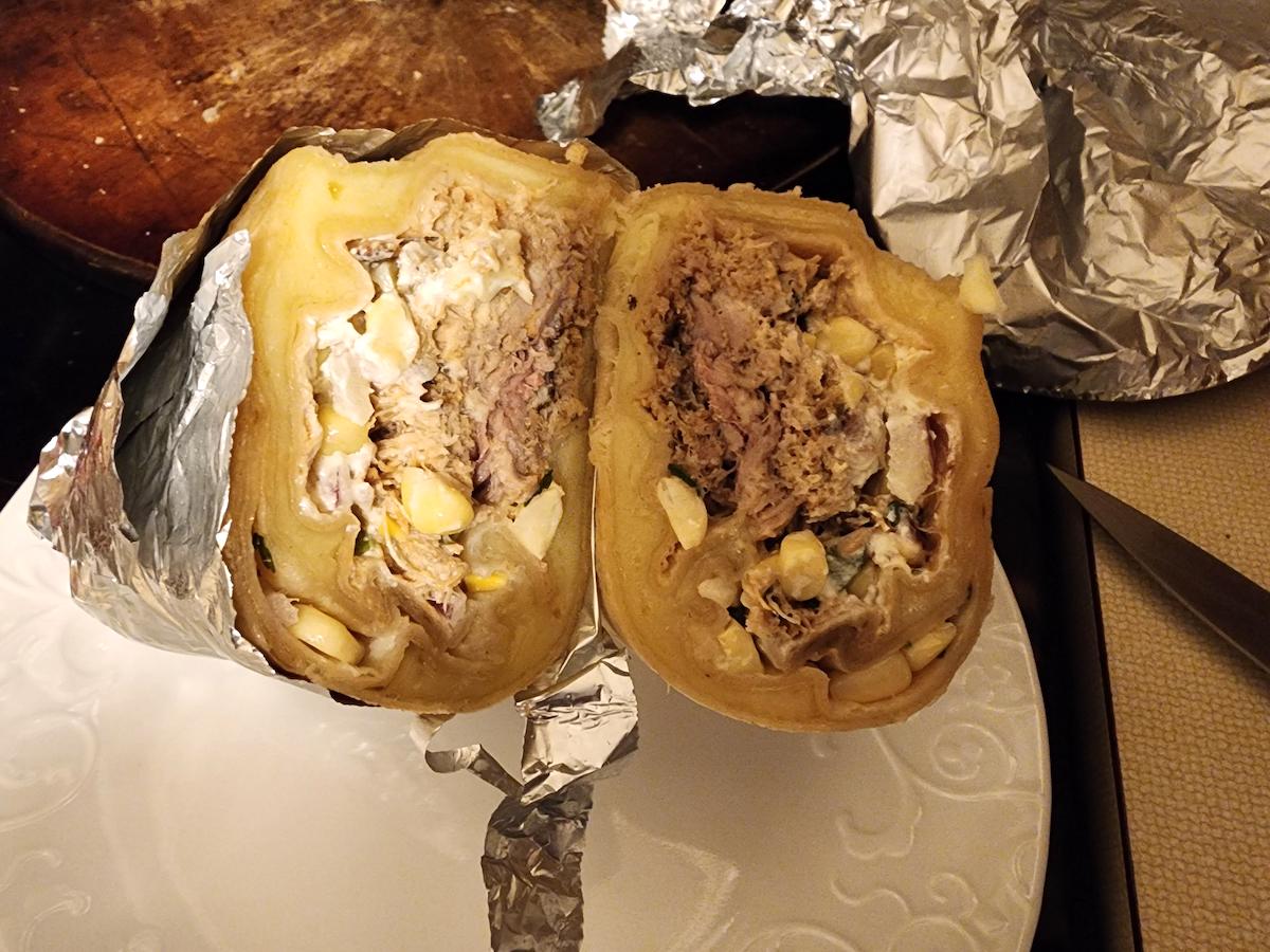 A foil-wrapped burrito cut in half on a white plate, showing a filling of shredded meat, corn, creamy sauce, and other ingredients. A knife and more foil are visible nearby.
