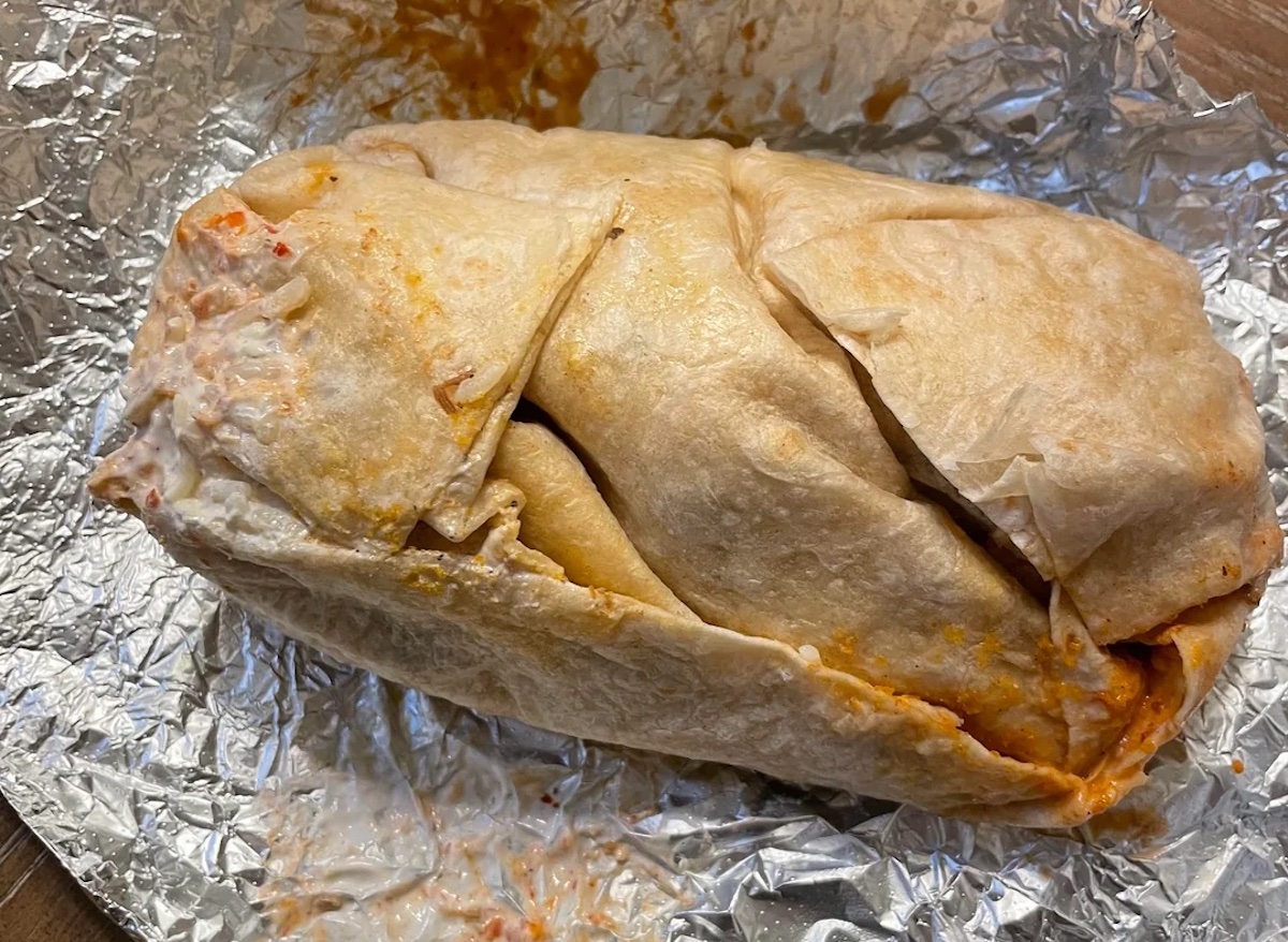 A large, wrapped burrito sits on crinkled aluminum foil. The tortilla is slightly torn and some creamy orange sauce is visible on the burrito and foil.