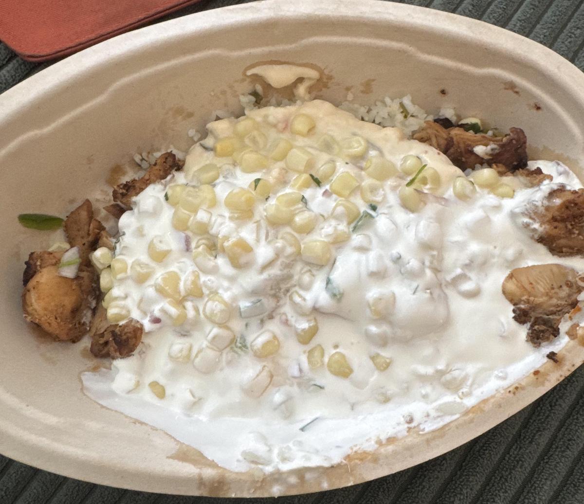 An oval bowl filled with grilled chicken pieces, white rice, corn kernels, and a generous amount of white sauce, possibly sour cream or crema, on top. The dish appears to be a burrito bowl.
