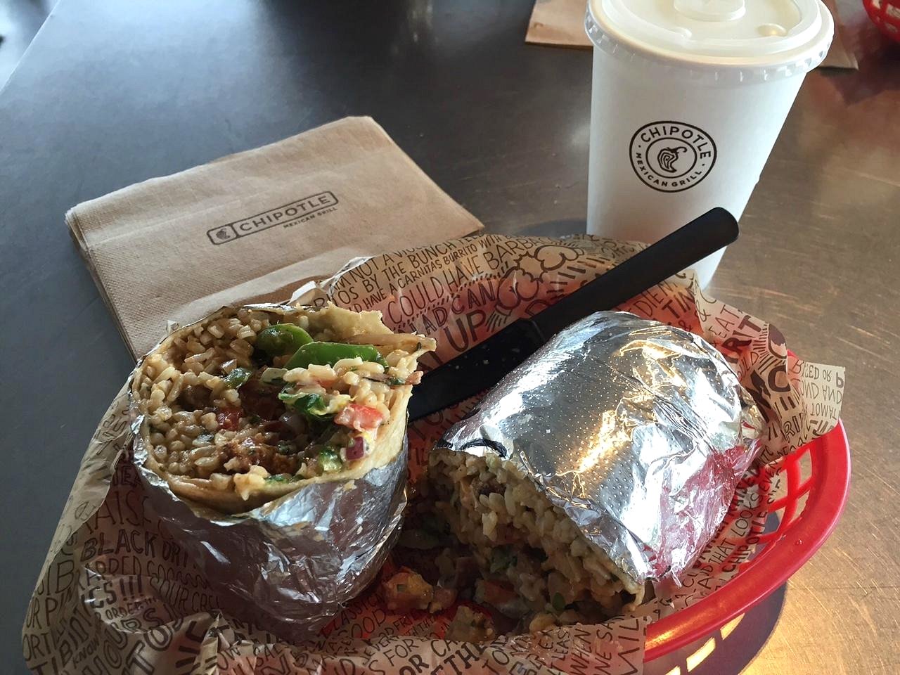 A burrito cut in half, wrapped in foil, sits on Chipotle-branded paper with a black plastic knife. A Chipotle drink cup and napkin are in the background on a metal table.