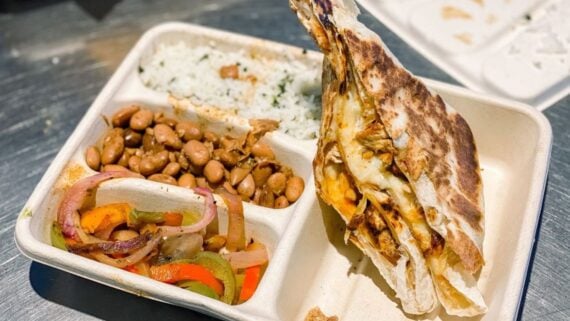 A takeout container with white rice, pinto beans, sautéed onions and peppers, and two cheesy quesadilla slices with shredded meat on a metal surface.