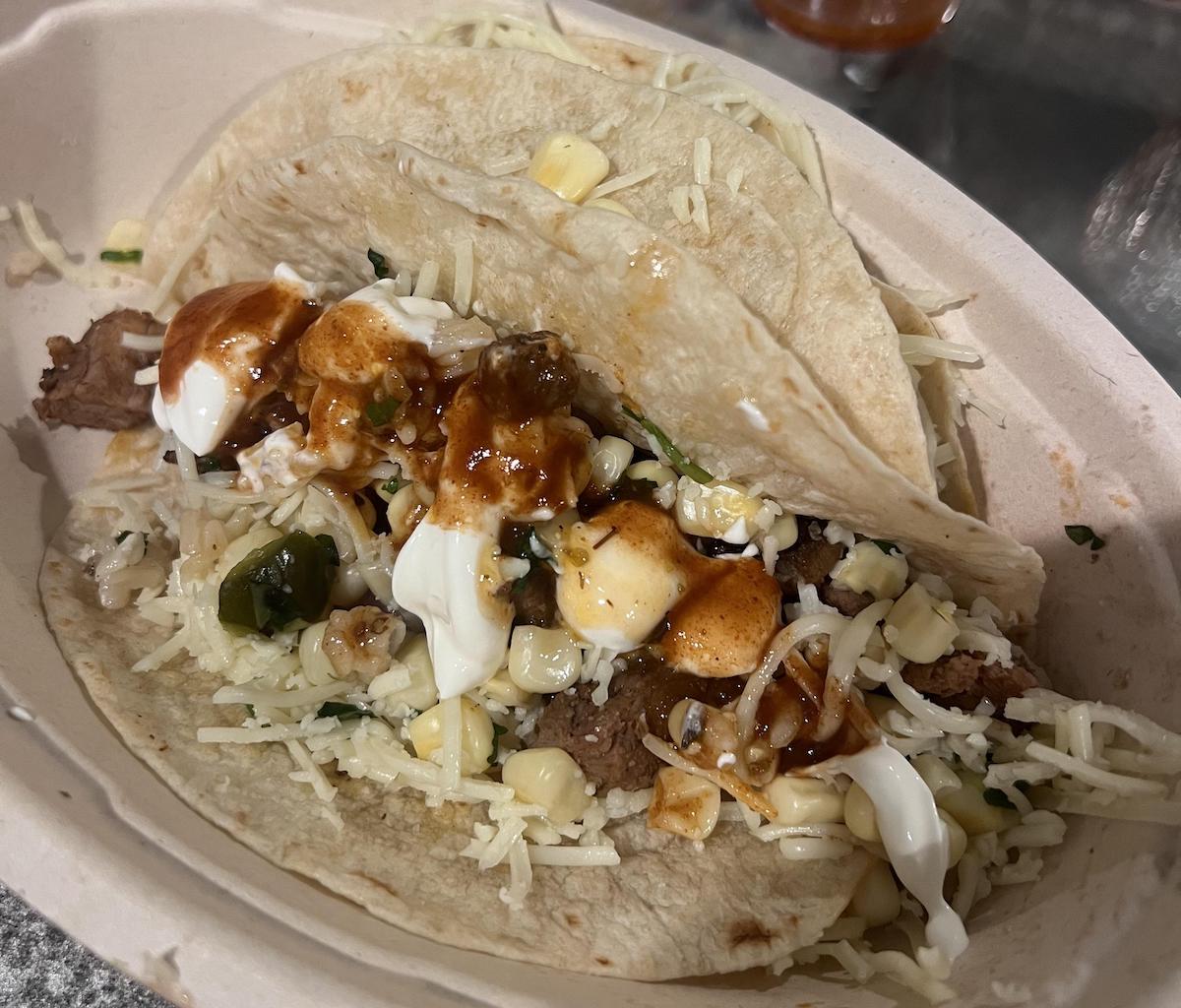 A close-up of a taco with grilled beef, corn, shredded cheese, sour cream, and brown sauce in a soft flour tortilla, served in a takeout container.