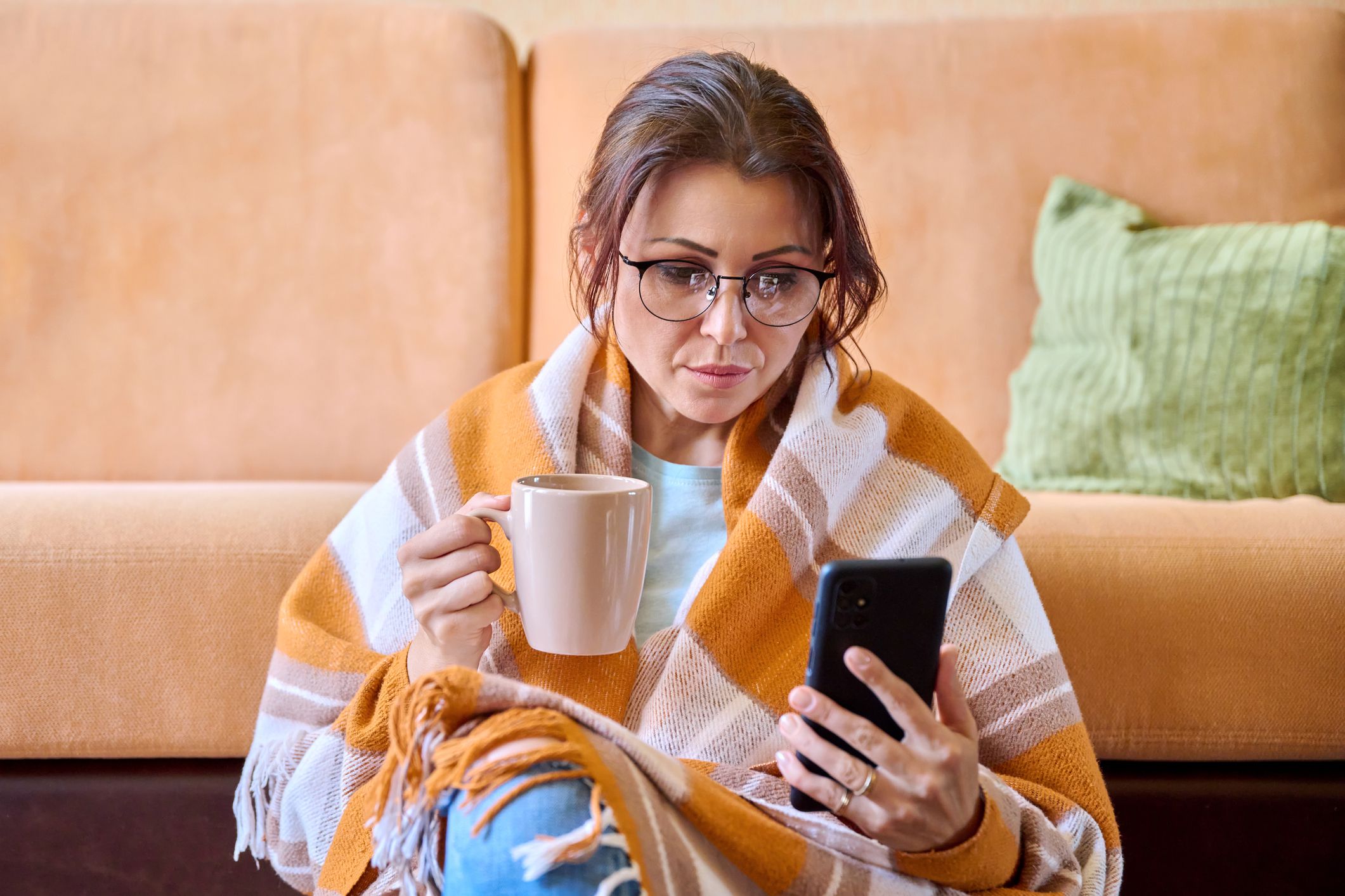 The season of cold weather is autumn winter, a woman under a woolen warm blanket with a cup of hot tea using a smartphone. Middle-aged female resting relaxing at home