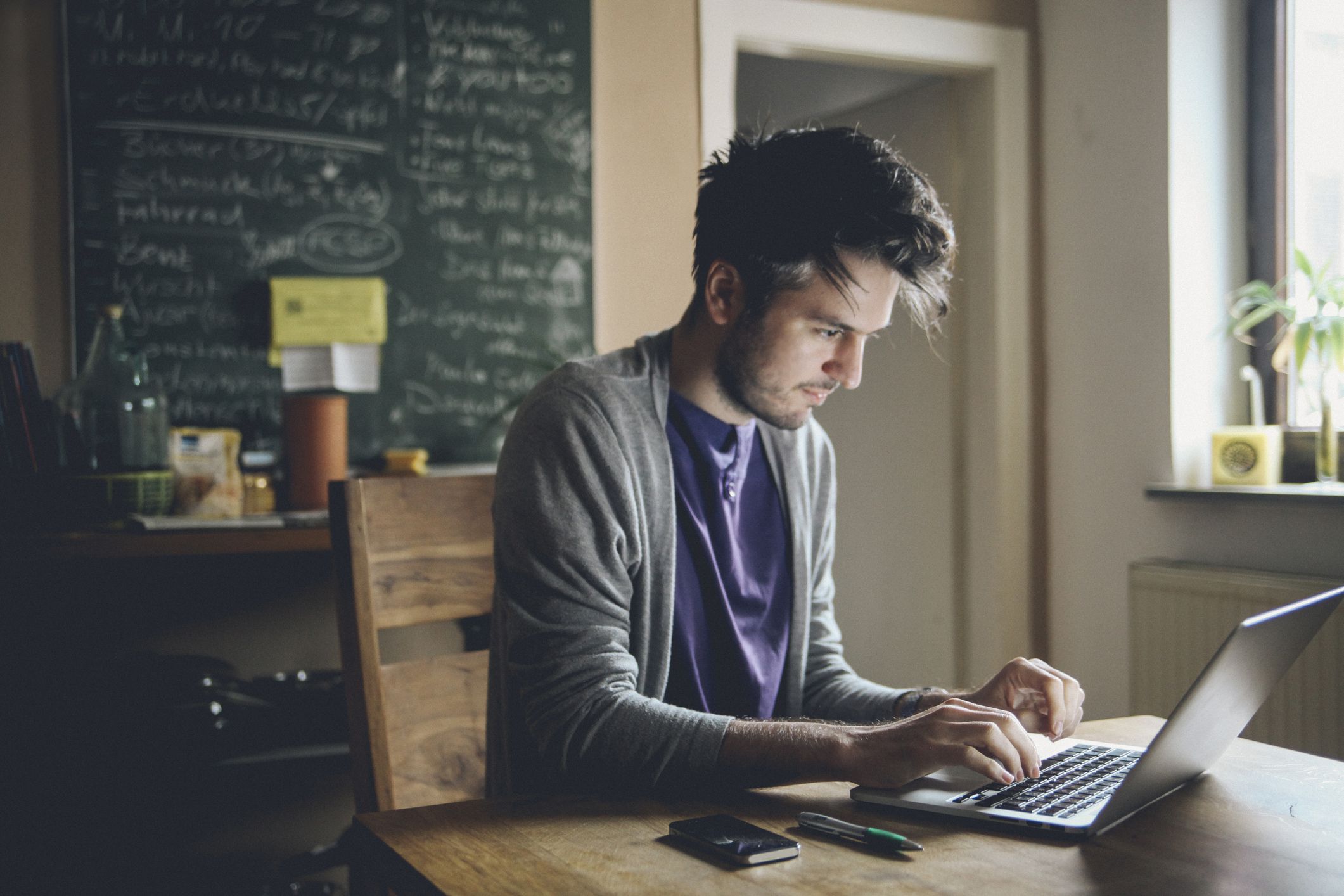 Vintage toned image of a young man working at home, freelancer, designer or writer, checking e-mails or typing on the laptop. Only natural light used, higher iso settings. Interior looks like a modern office, home or a co-working space.