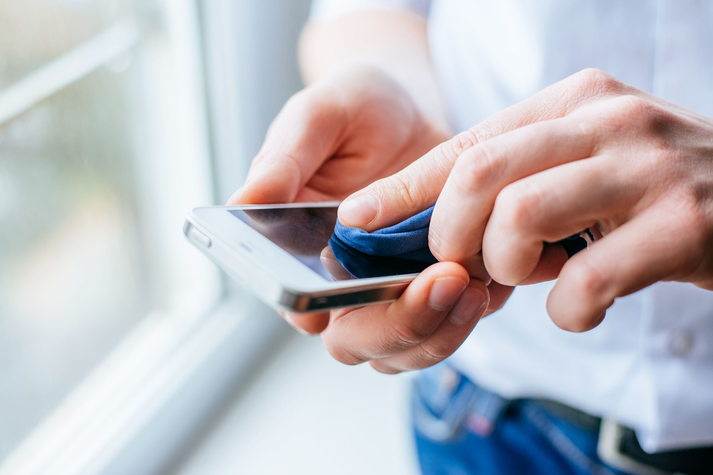 Closeup of two male hands cleaning a smartphone while holding it with a blurred background of the midsection of male and a room window