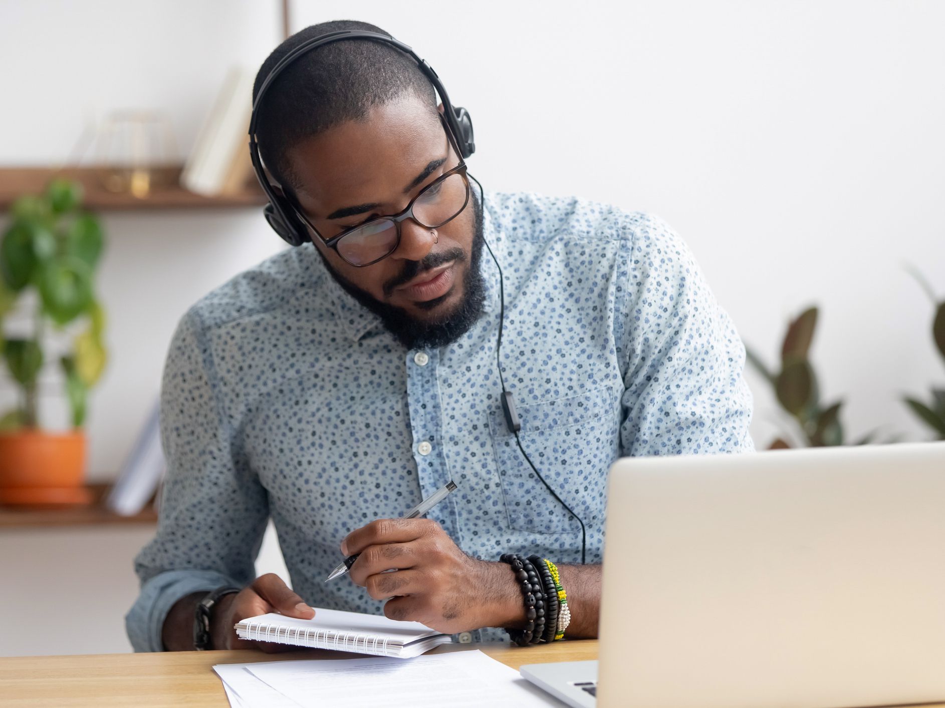 Focused african business man in headphones writing notes in notebook watching webinar video course, serious black male student looking at laptop listening lecture study online on computer e learning