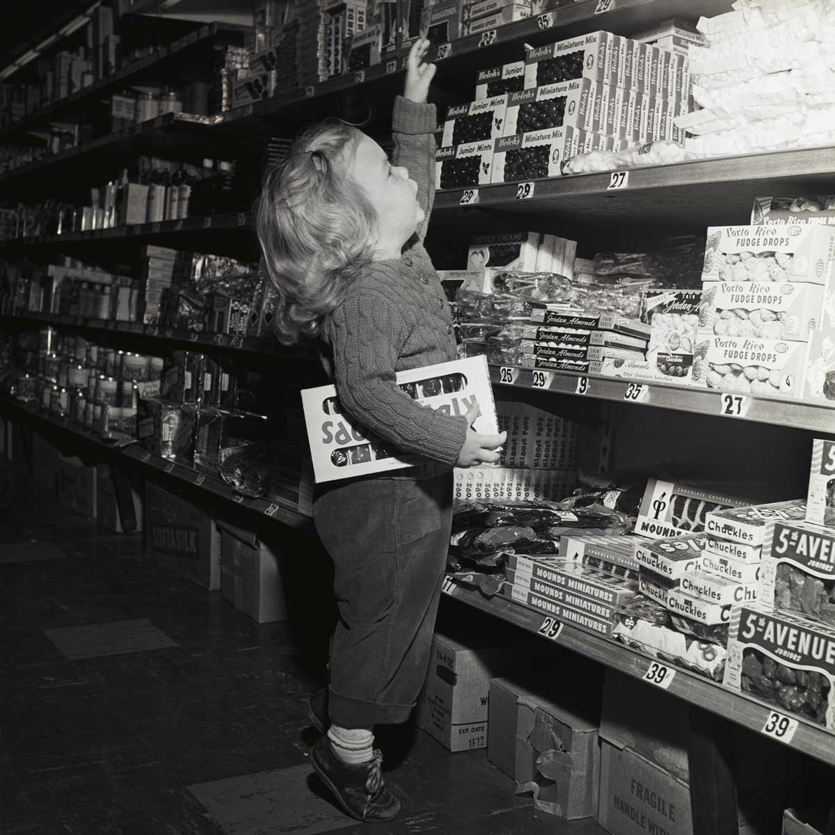 A young child stands in a grocery store aisle, reaching up toward a high shelf stocked with candy bars while holding a box in their arm. The shelves are filled with various snacks and sweets.
