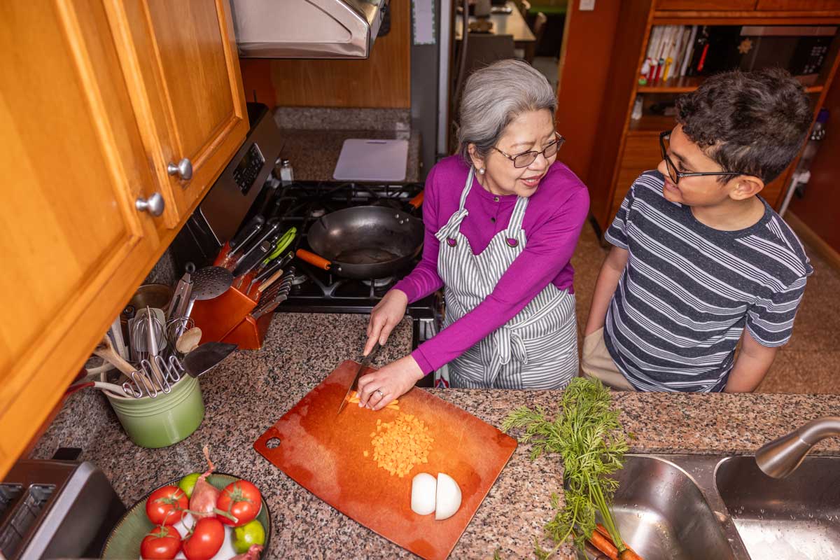 An older woman in an apron chops carrots on a cutting board in a kitchen while smiling and talking to a young boy standing beside her. Fresh vegetables are on the counter near them.