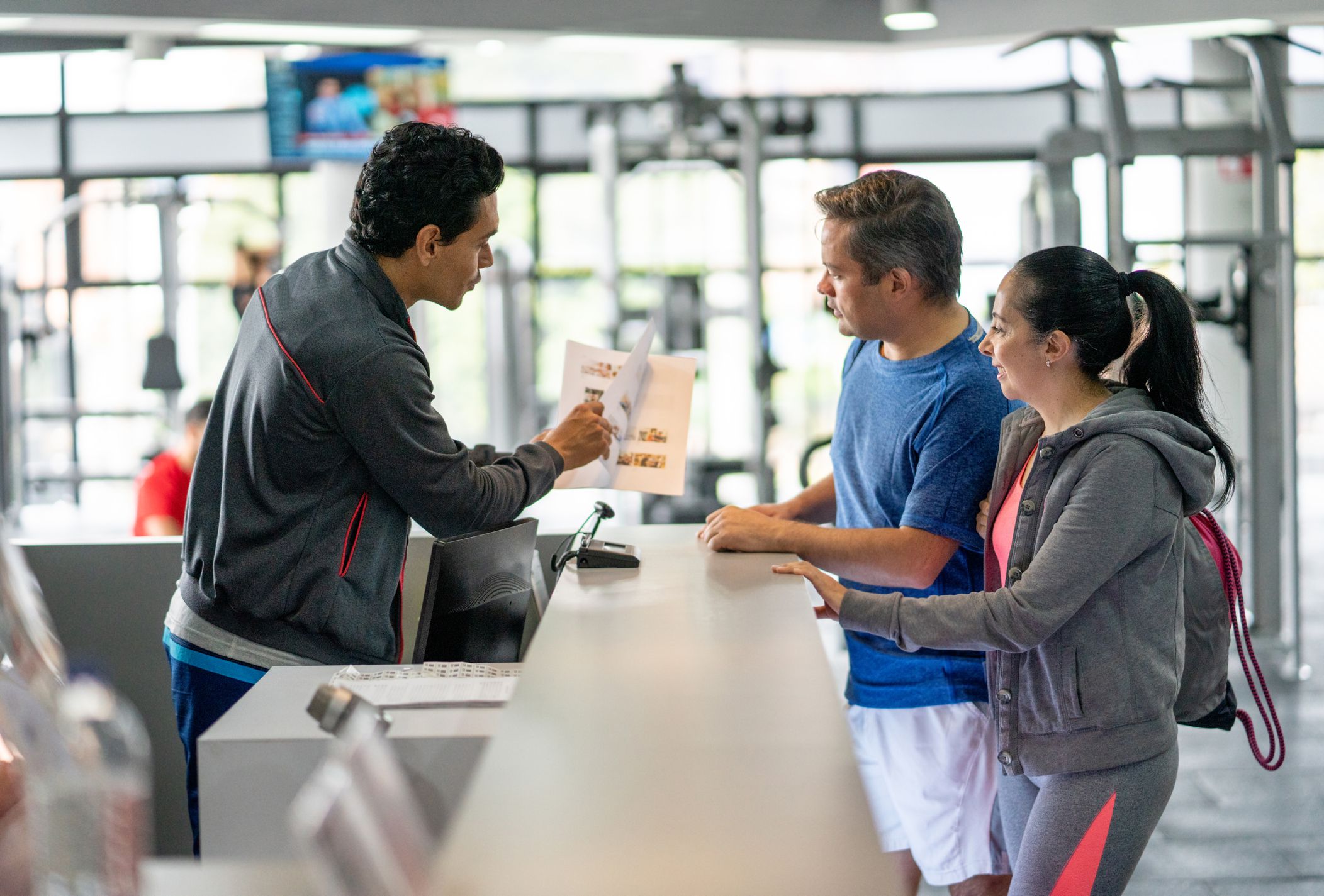 Happy couple registering at the gym while employee explains benefits showing them a catalogue - healthy lifestyles