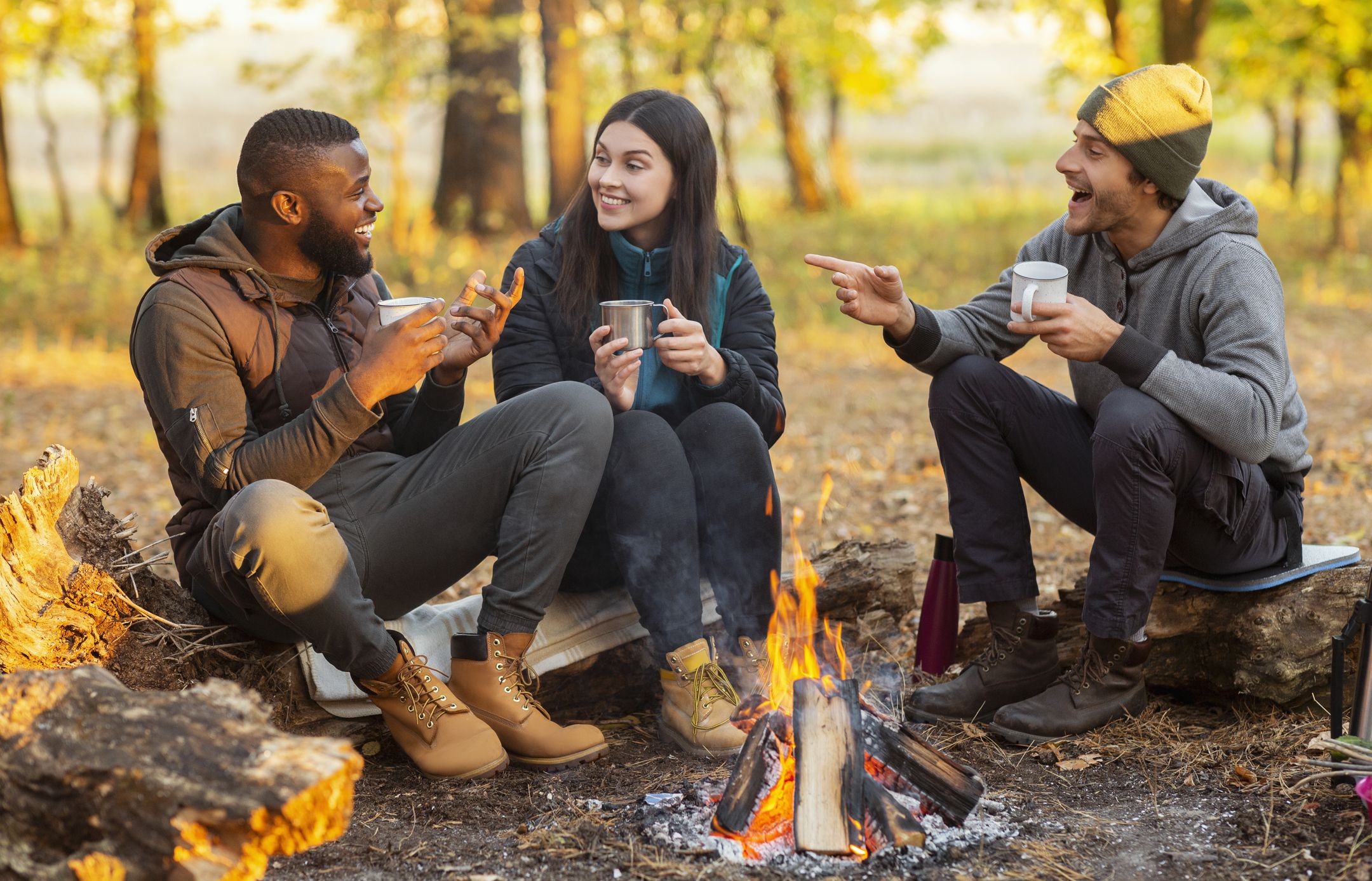 Camping time for mixed race group of friends sitting beside fireplace and frying sausages in autumn forest in sun lights
