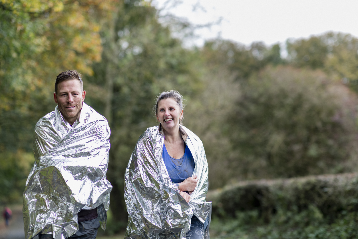 A man and woman walking outdoors, wrapped in silver thermal blankets, smiling and appearing cheerful with a blurred background of green trees and foliage.