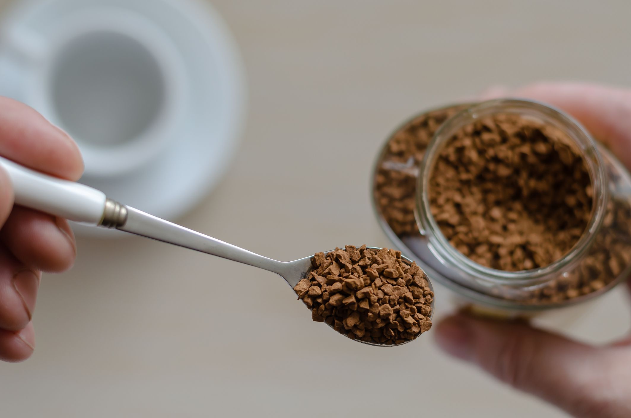Glass jar with coffee in a hand. A hand holds a spoon full of instant coffee. Pile of instant coffee grains in metal spoon over glass jar. Caffeine drink. View from above. Selective focus. Close-up.