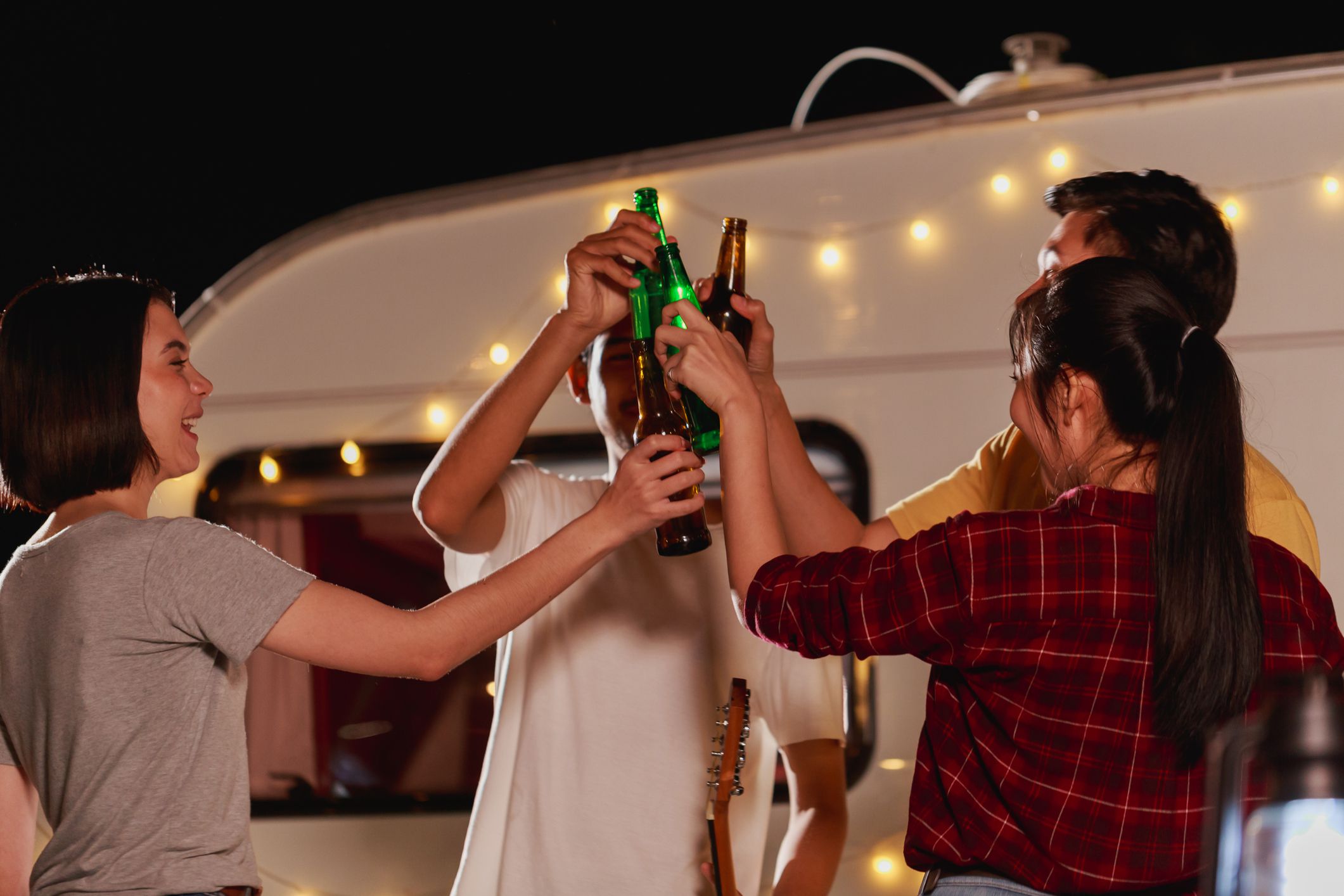 Group of young diverse friends holding the bottle drinking talking and enjoying a picnic in the park in the evening playing music having fun on the camping area by the nature.