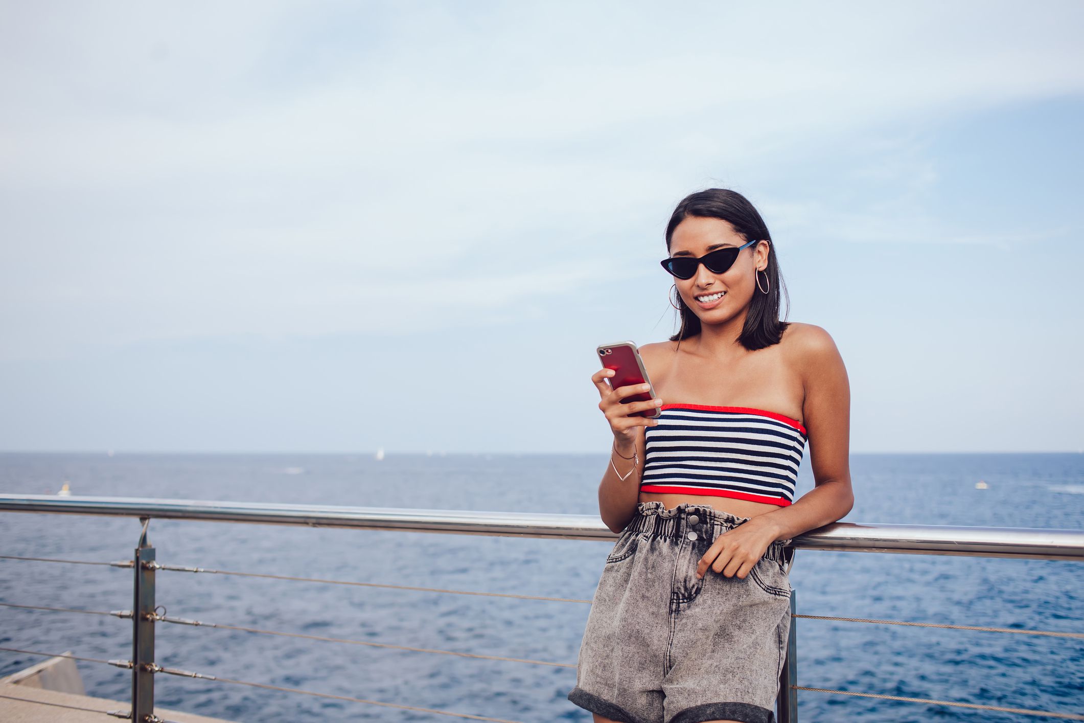 Optimistic female tourist in stylish clothing standing at seafront and chatting online via cellphone while chilling during summer vacation in Rome