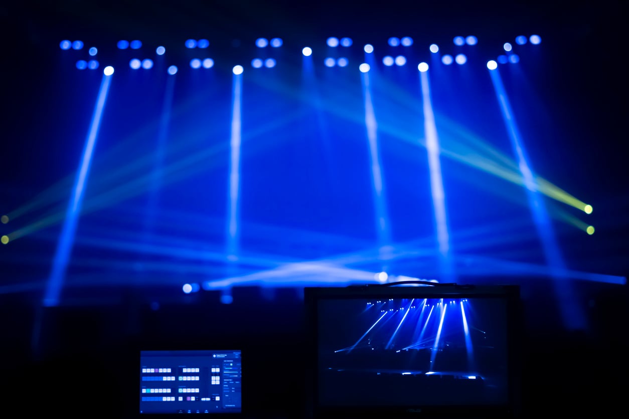 A stage with blue and yellow spotlights shining through haze, viewed from behind a control booth with two screens displaying lighting controls and the stage scene.