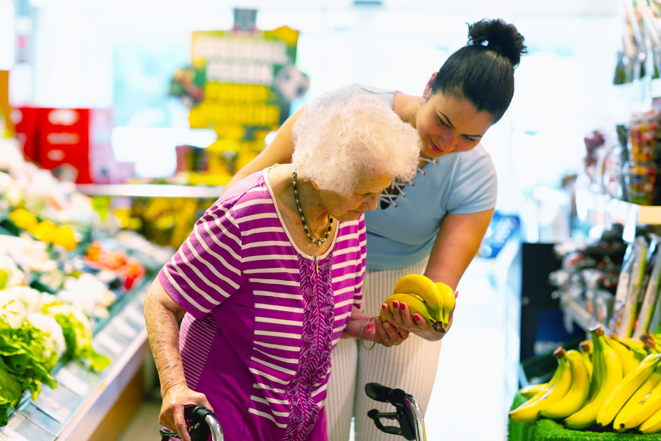 Young hispanic caregiver supporting senior woman, 93 years, with buying banans in a shop