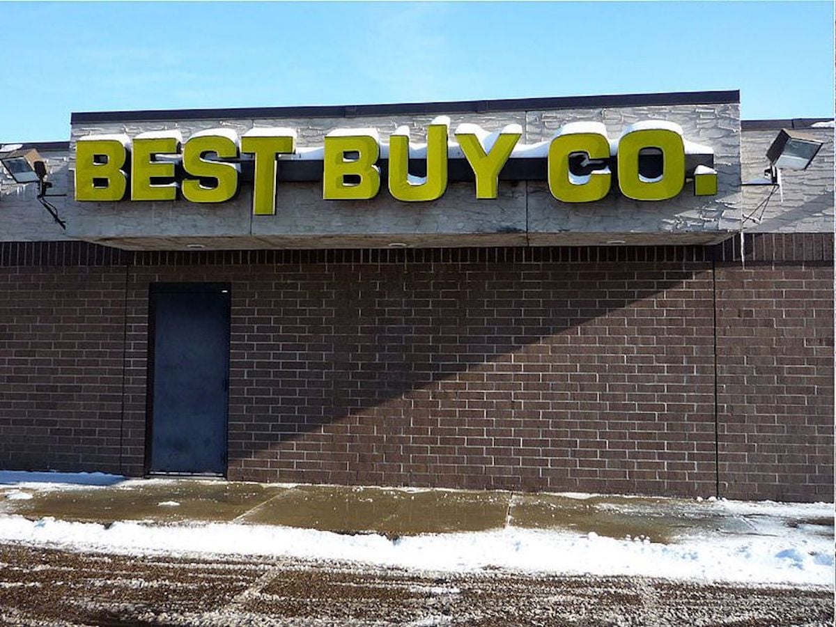 A brick building with a large yellow "BEST BUY CO." sign above a black door, representing one of the notable companies founded by year. Snow is piled along the sidewalk and edge of the building, and the sky is clear and blue.