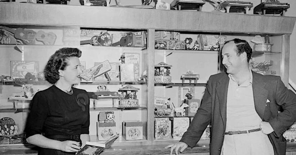 A woman and a man stand in front of a display case filled with vintage toys and decorative items from companies founded by year. The woman holds an object, smiling, while the man leans casually against the case, engaged in conversation.