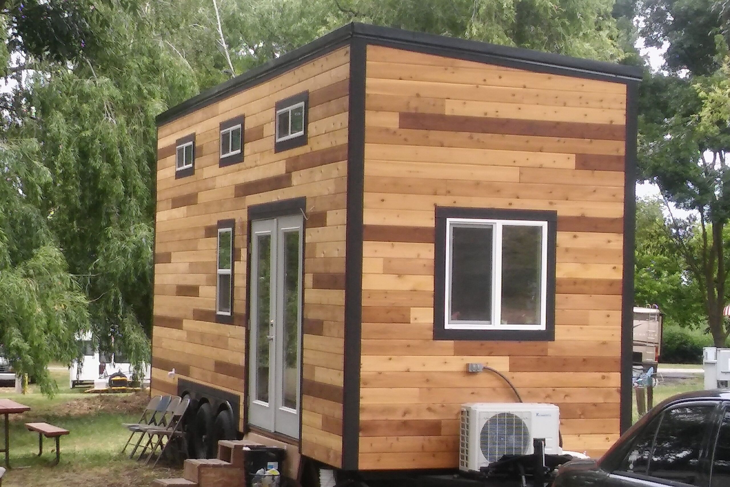 A tiny home in Park Delta Bay, Isleton, California, wooden exterior, surrounded by lush trees, an RV, cars, and another tiny home in the background