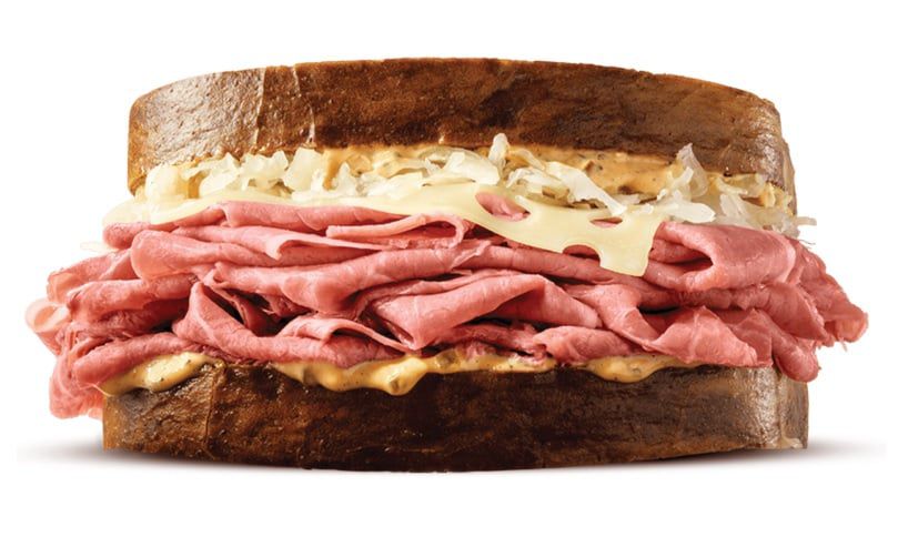A close-up of a sandwich with layers of sliced roast beef, melted Swiss cheese, sauerkraut, and sauce on dark rye bread, against a white background.