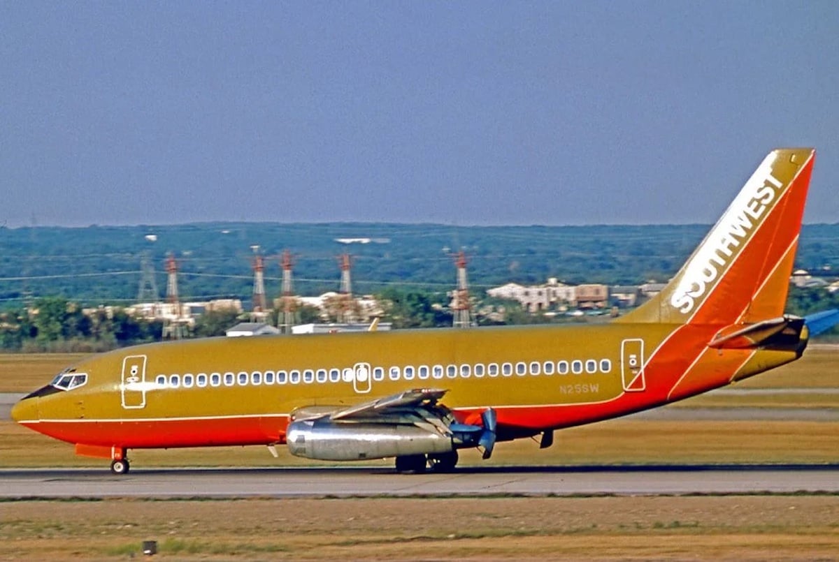 A vintage Southwest Airlines jet, from one of the notable companies founded by year 1967, with a gold, red, and orange livery taxis on a runway, with buildings and trees visible in the distant background.