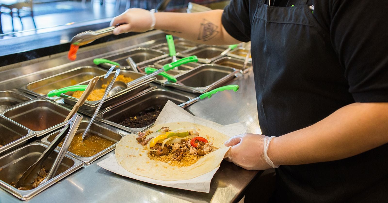 A person wearing gloves prepares a burrito, adding ingredients like rice, shredded meat, and peppers from a selection of metal containers at a food counter.