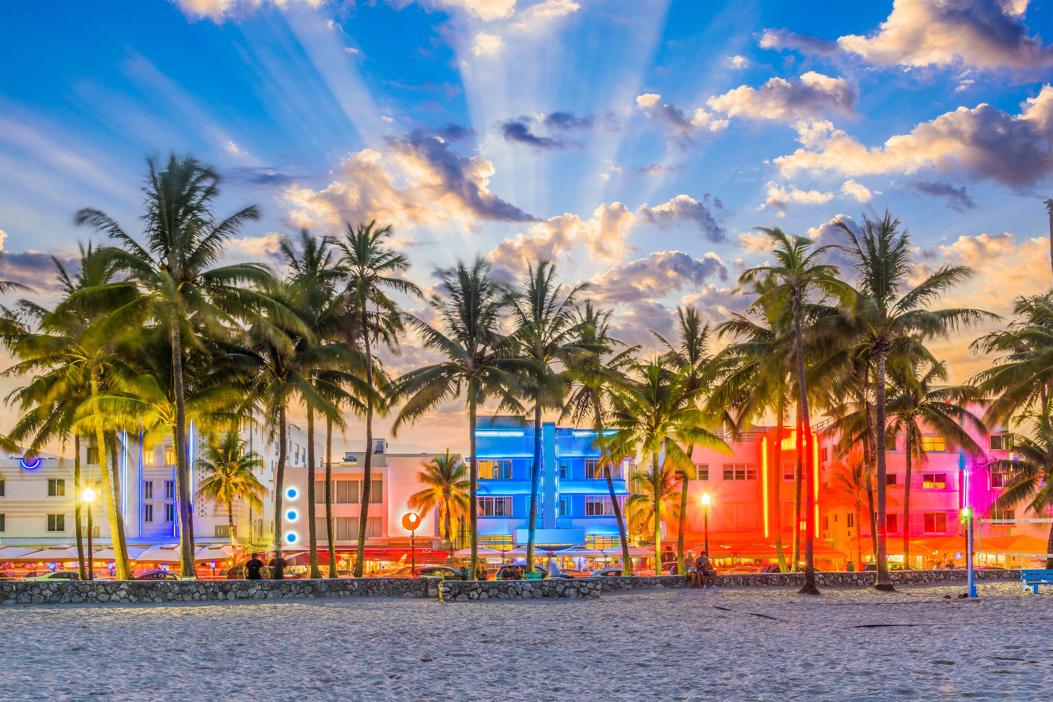 Dramatic lights from buildings along Ocean Drive, Miami Beach, Florida during early sunset, beach in the foreground with a line of palm trees, blue, yellow, and red lights