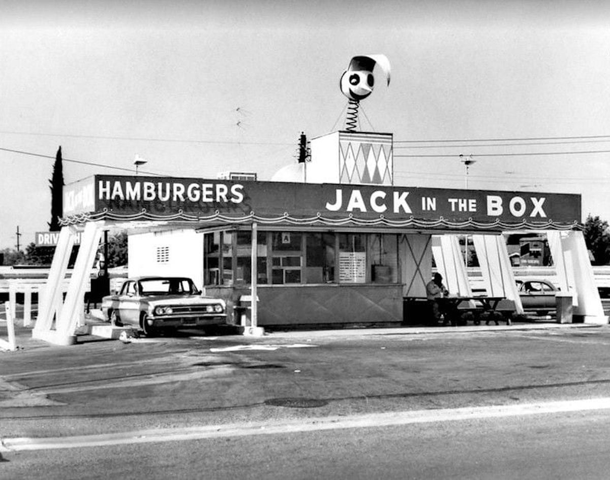 Black-and-white photo of a vintage Jack in the Box fast food restaurant, one of the iconic companies founded by year, with a large Jack figure on the roof, a classic car at the drive-thru, and a person sitting at an outdoor table.