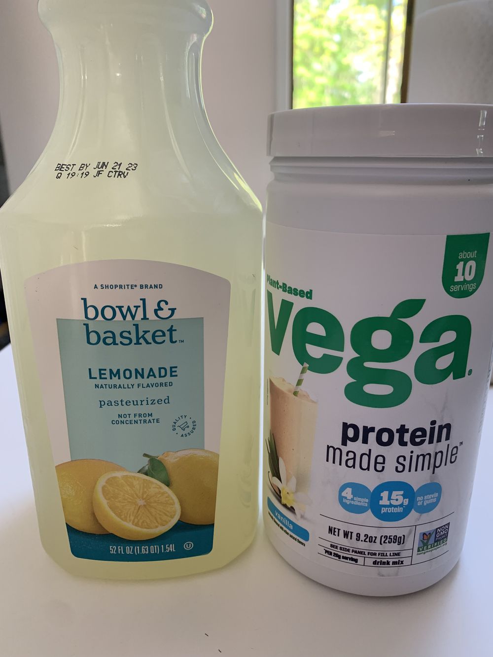 A bottle of Bowl & Basket lemonade and a container of Vega plant-based protein powder are placed side by side on a white surface. The labels are clearly visible.
