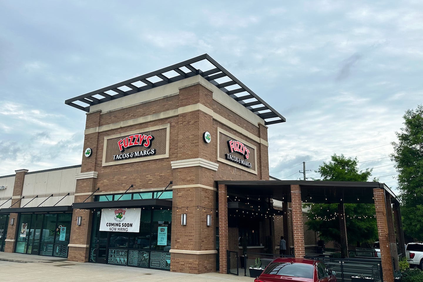 A brick restaurant with signs reading "Fuzzy's Tacos & Margs." A banner on the window says "Coming Soon." There is a covered outdoor seating area, some greenery, and a red car parked in front. The sky is cloudy.