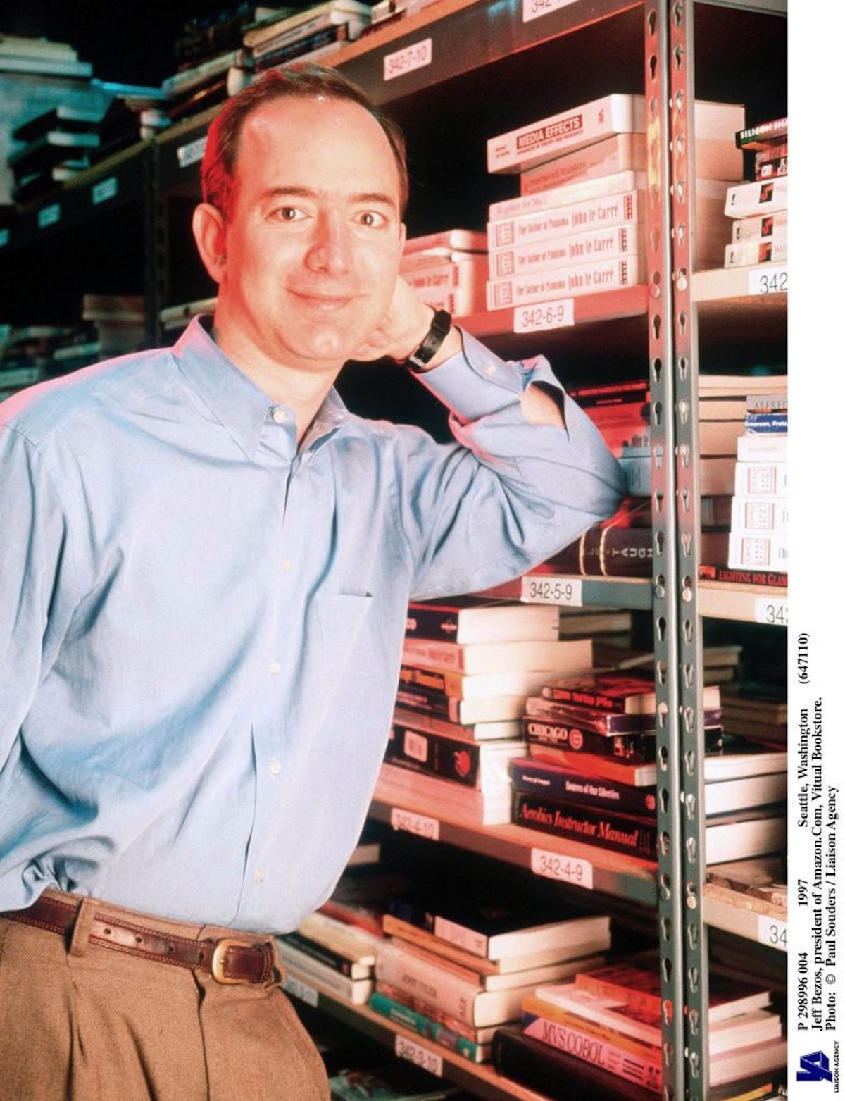 A man in a blue shirt leans on a metal shelf filled with books and VHS tapes, smiling at the camera in what appears to be a storage room, perhaps organizing records of companies founded by year.